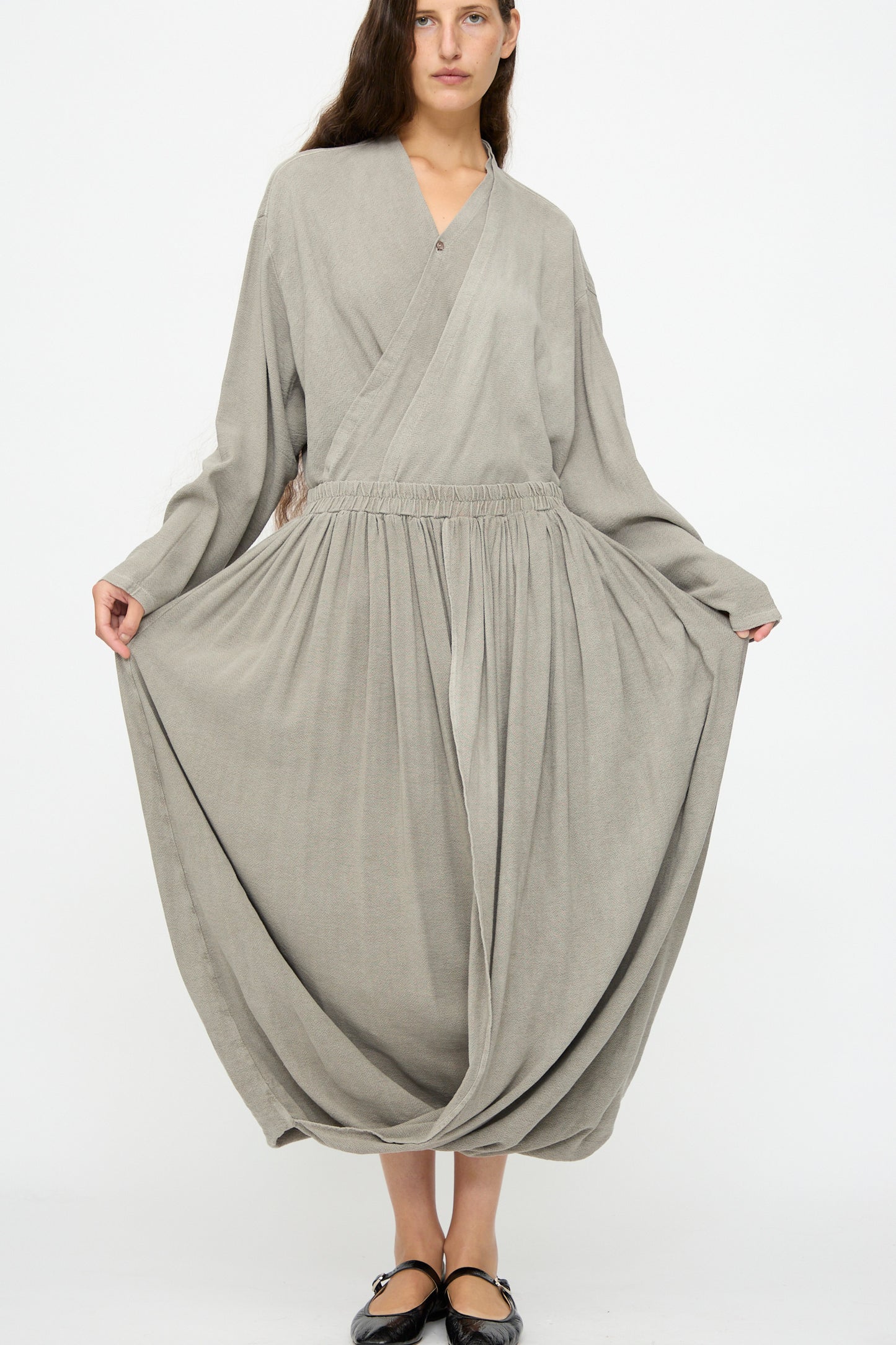 A woman stands against a white background wearing the Black Crane Double Layered Drapy Skirt in Dark Stone—a loose-fitting, midi-length skirt with an elastic waistband—paired with a long-sleeved top.