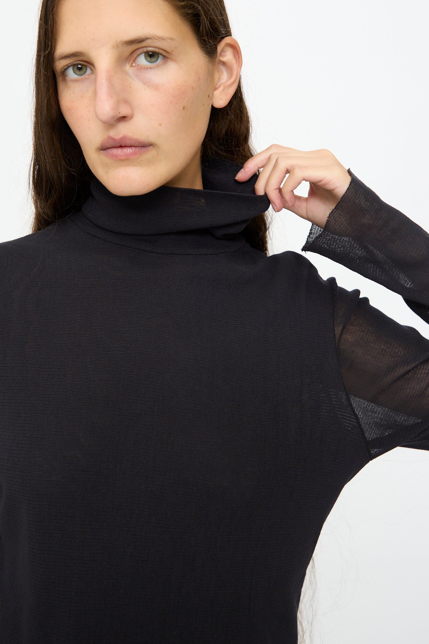 A person with long brown hair wears the Black Crane Double Mesh Turtleneck in Black, holding the collar with one hand and looking directly at the camera against a plain background.