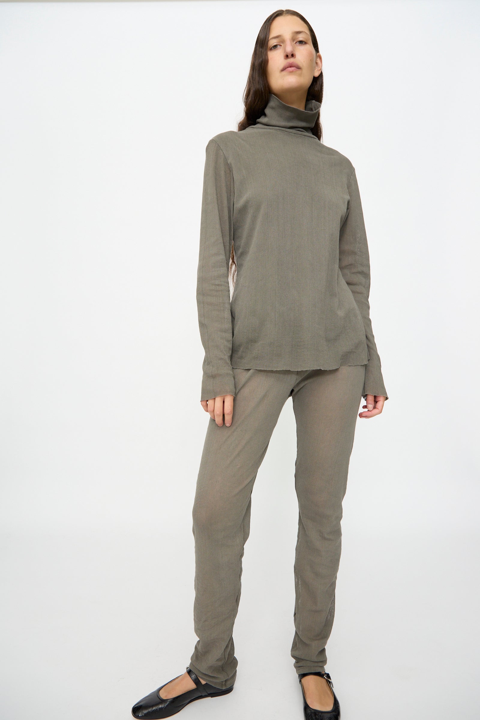 A person stands in a studio wearing the Black Crane Double Mesh Turtleneck in Dark Stone with matching pants and black flat shoes against a plain white background.