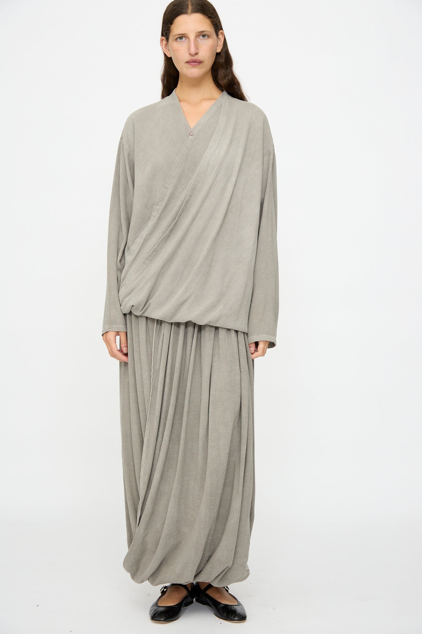A person stands against a plain white background, wearing the Black Crane Drapy Shirt in Dark Stone with a draped front skirt and black flats.