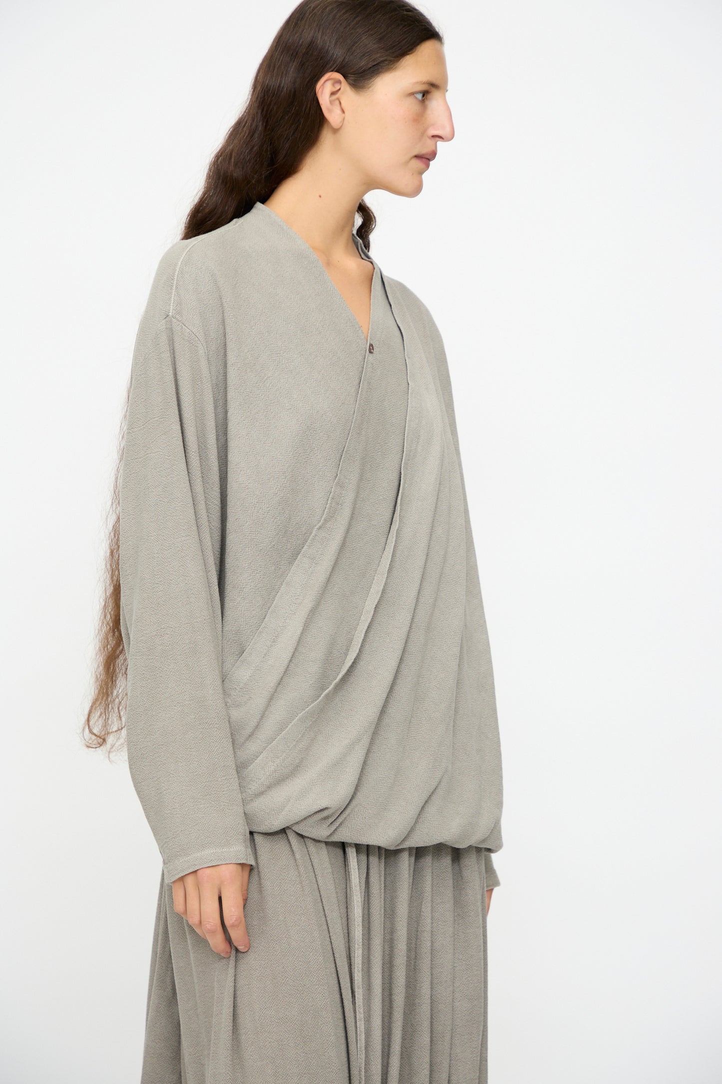 A person with long brown hair wears the Black Crane Drapy Shirt in Dark Stone, a loose-fitting top made from a soft cotton rayon blend, standing against a plain light background.
