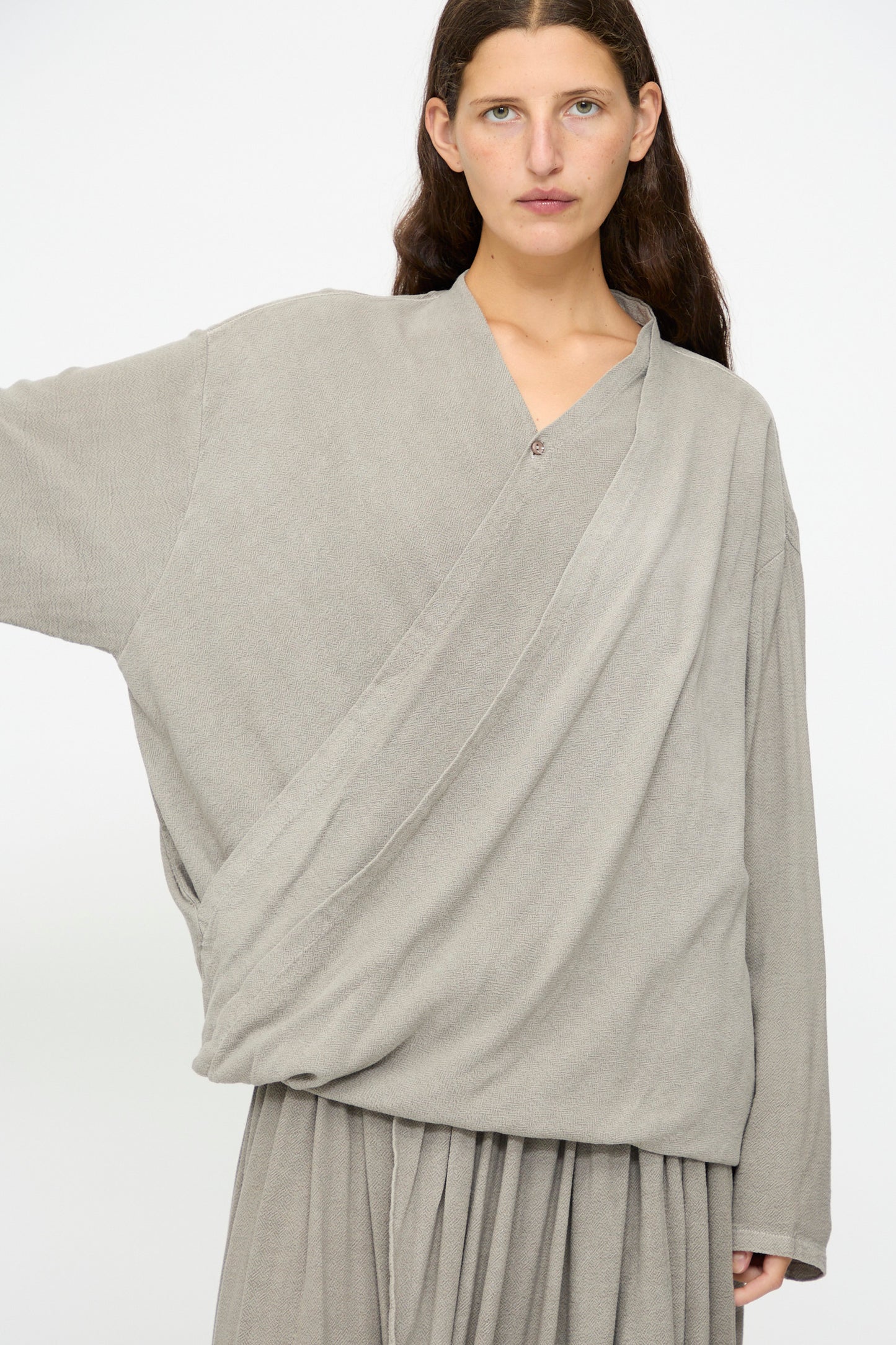 A person with long brown hair wears the Black Crane Drapy Shirt in Dark Stone, a loose-fitting cotton rayon blend top, paired with a matching skirt, standing against a plain white background.