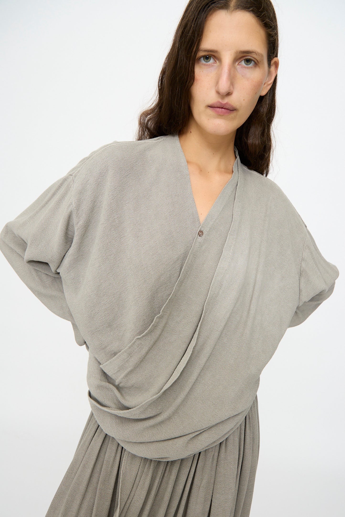 A person with long brown hair wears the Black Crane Drapy Shirt in Dark Stone, a loose-fitting long sleeve top with a draped front, paired with a matching light gray cotton-rayon skirt, standing against a plain background.