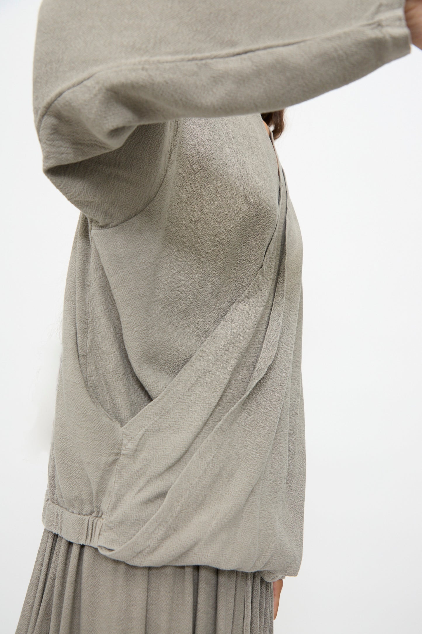 A person wearing the Black Crane Drapy Shirt in Dark Stone, a loose cotton-rayon blend with a draped front and long sleeves, is shown from the side raising their arm against a plain background.