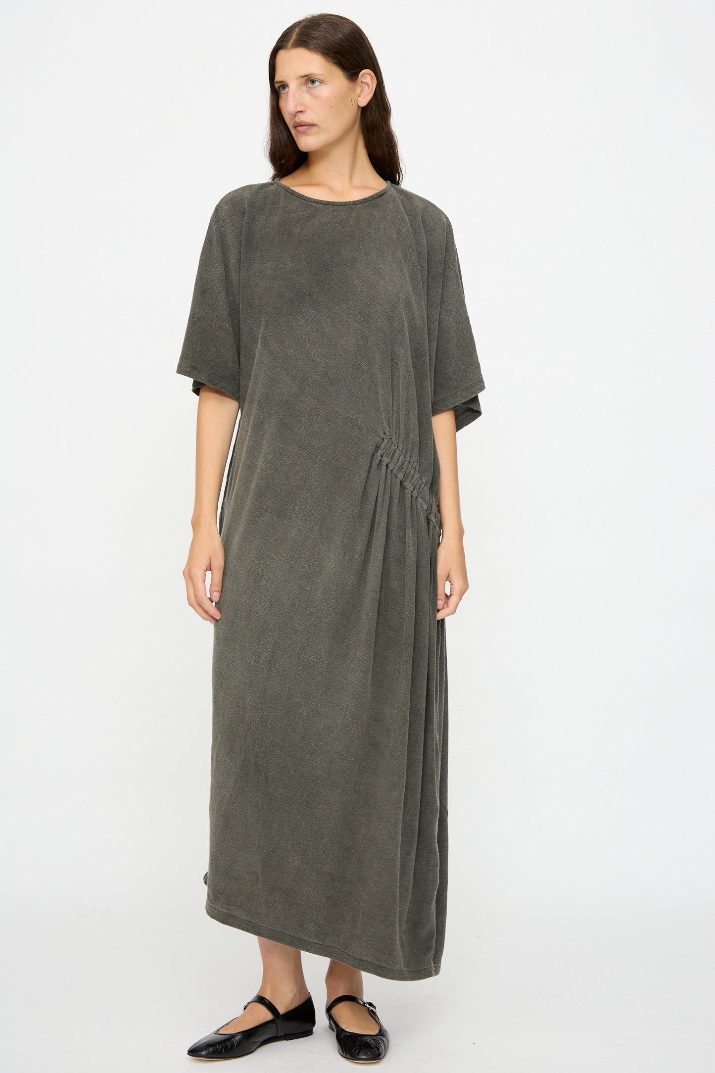 A person wears the Black Crane Greek Dress in Slate—a relaxed fit, ankle-length, short-sleeved maxi dress in a soft cotton-rayon blend with gathered side detailing—standing against a plain white background.