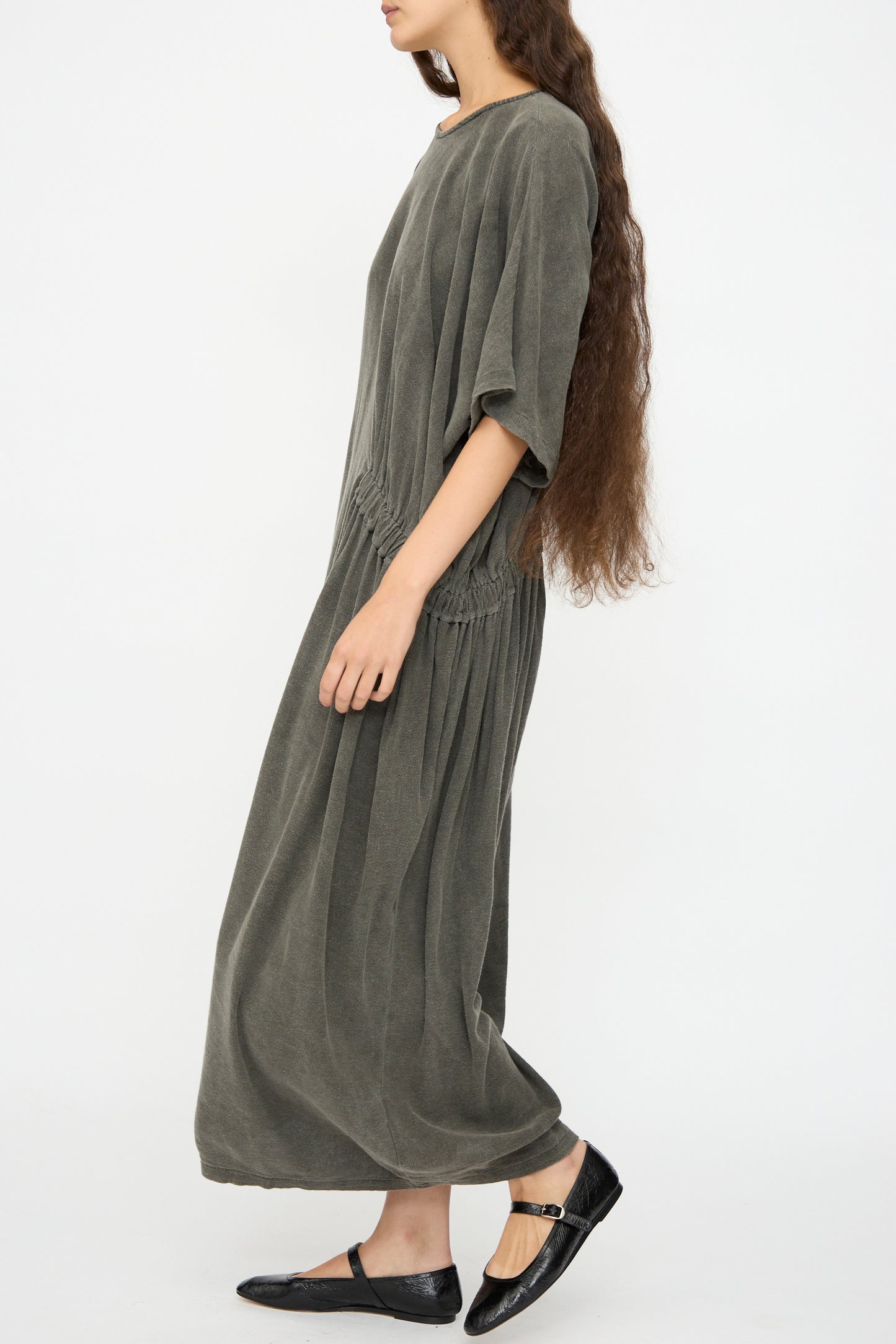 A woman with long, wavy brown hair wears the Black Crane Greek Dress in Slate, a relaxed fit maxi dress in a soft cotton rayon blend, paired with black flat shoes, standing against a plain white background.