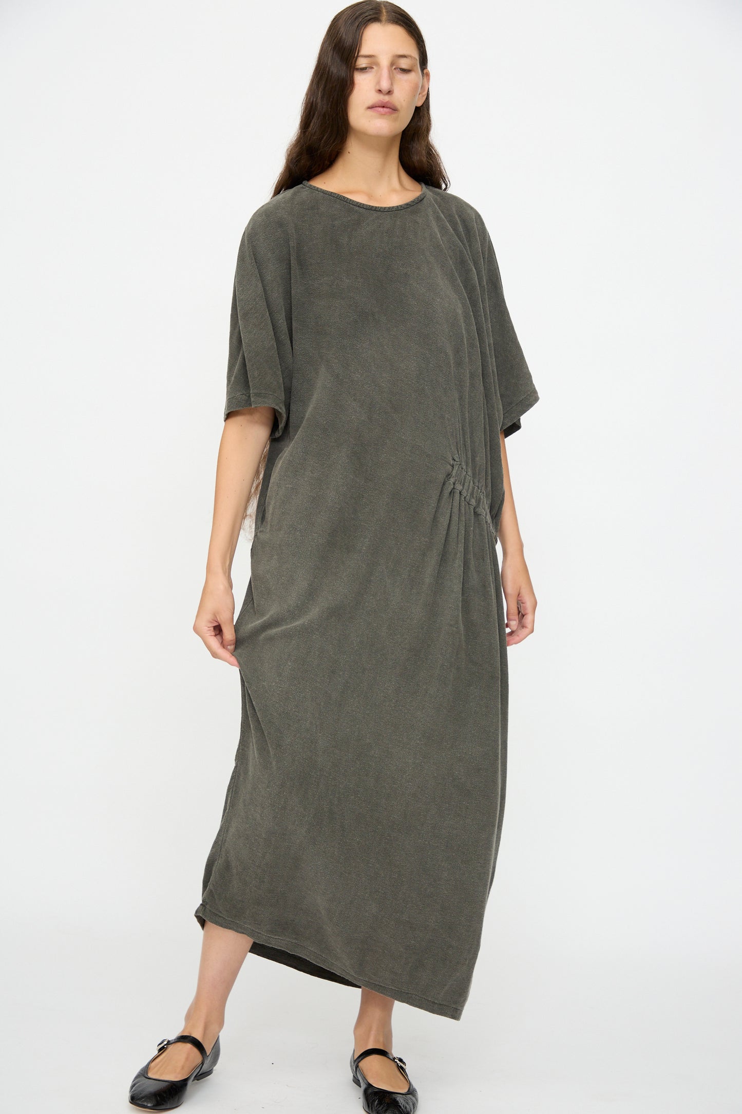 A person wears the Black Crane Greek Dress in Slate, a relaxed fit, dark gray maxi made from a soft cotton-rayon blend, paired with black flats against a plain background.