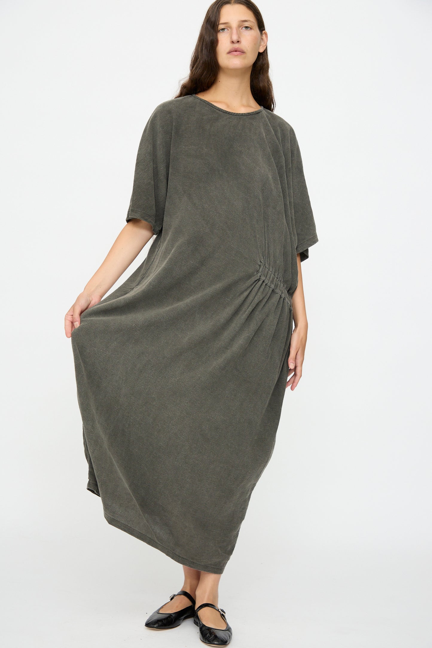 A person wears the Black Crane Greek Dress in Slate, a relaxed-fit, dark gray maxi dress with short sleeves and gathered side detail, made from a soft cotton-rayon blend, standing against a plain white background.