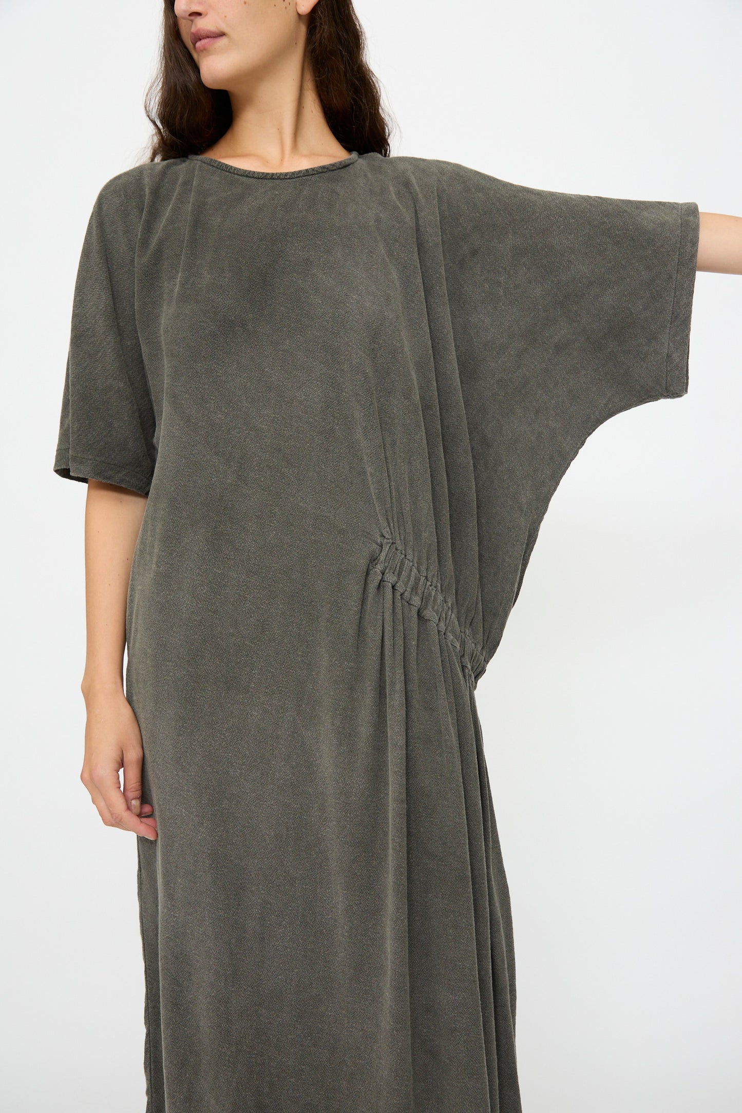 A person wears the Black Crane Greek Dress in Slate—a relaxed-fit, short-sleeved grey dress with waist gathers, made from a soft cotton-rayon blend—standing against a plain white background.