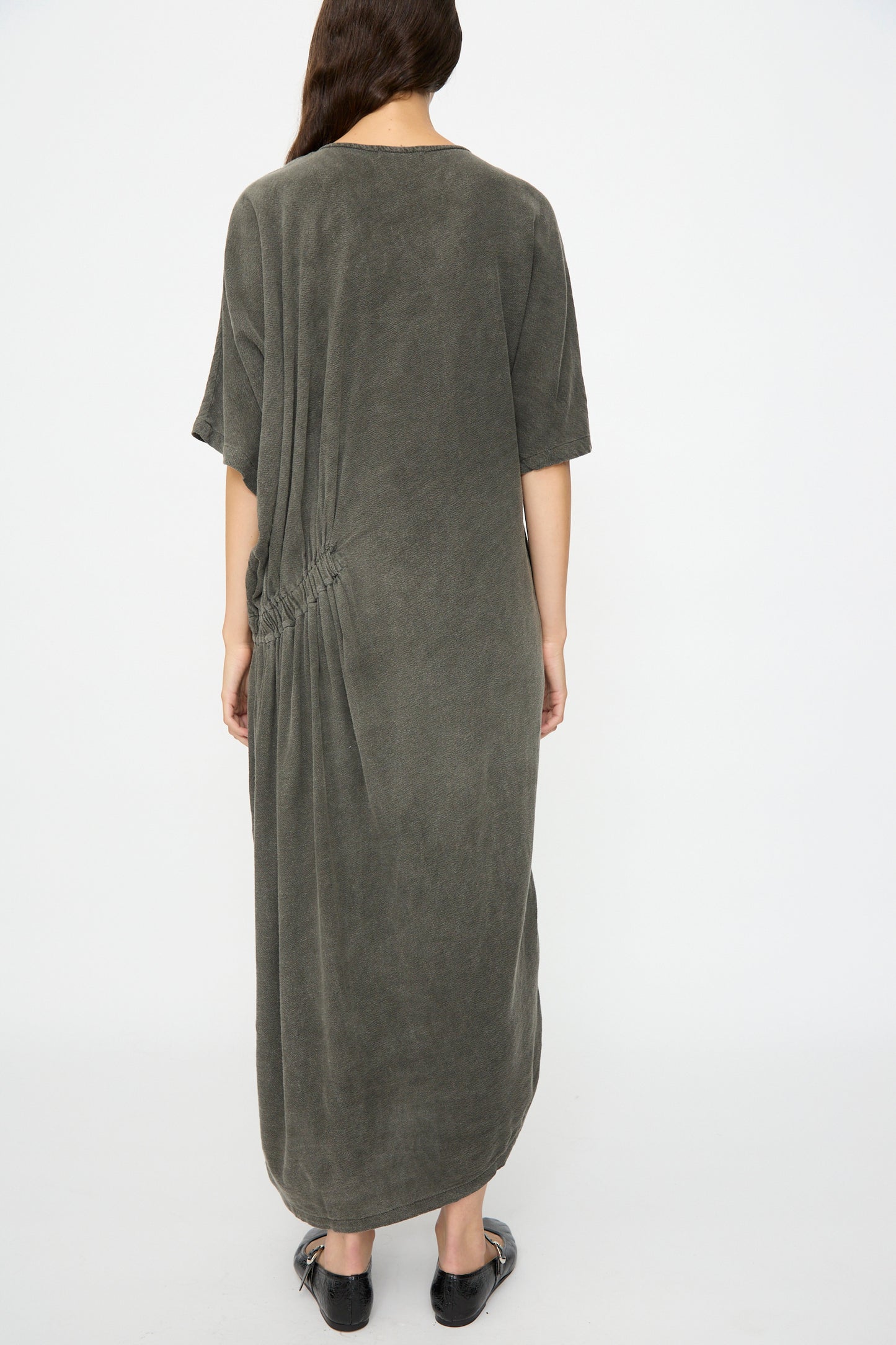 Person stands with back to camera wearing the Black Crane Greek Dress in Slate, a relaxed fit, ankle-length maxi with short sleeves and ruched side detail, paired with flat black shoes; hair over one shoulder.