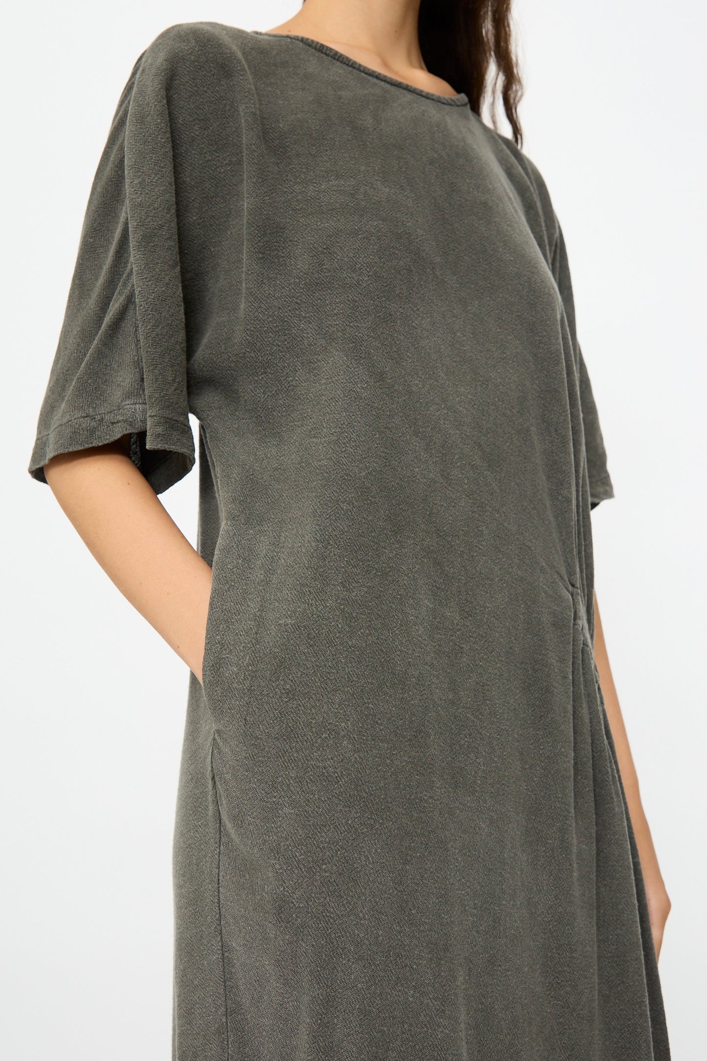 A person wears the Black Crane Greek Dress in Slate—a relaxed-fit, short-sleeve, dark gray cotton-rayon blend dress with side pockets—shown from shoulders to knees against a plain light background.