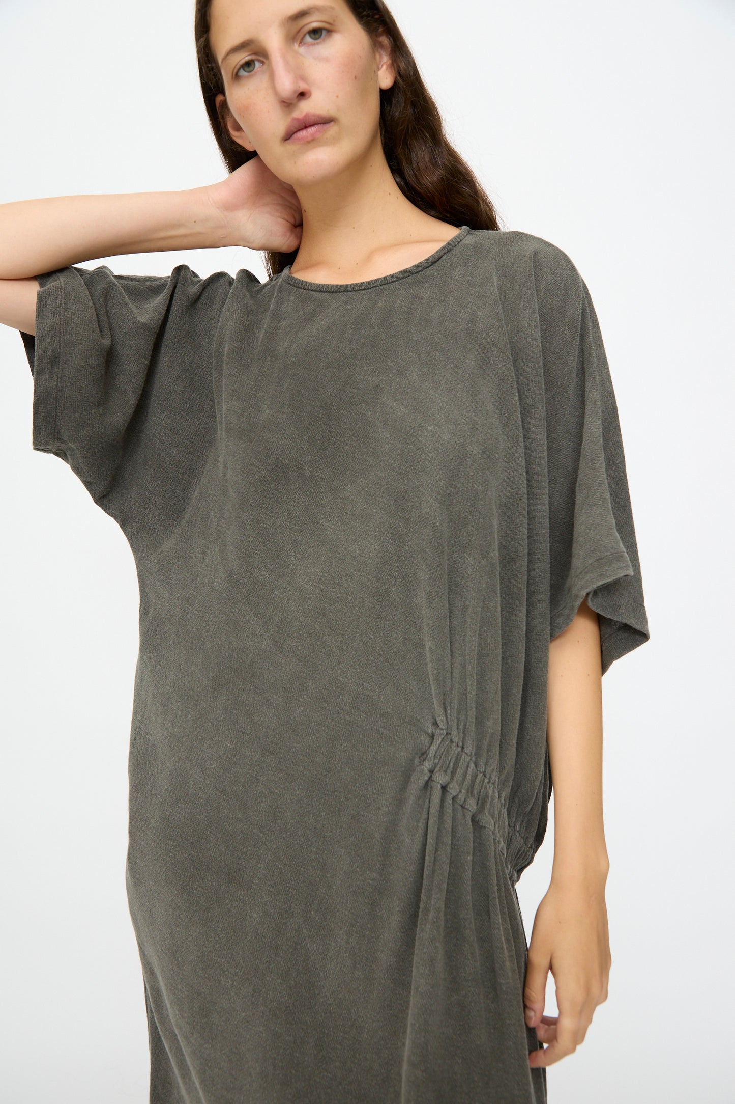 A person wears the Black Crane Greek Dress in Slate, a relaxed-fit, short-sleeved dark grey dress with waist ruching, standing against a plain light background.