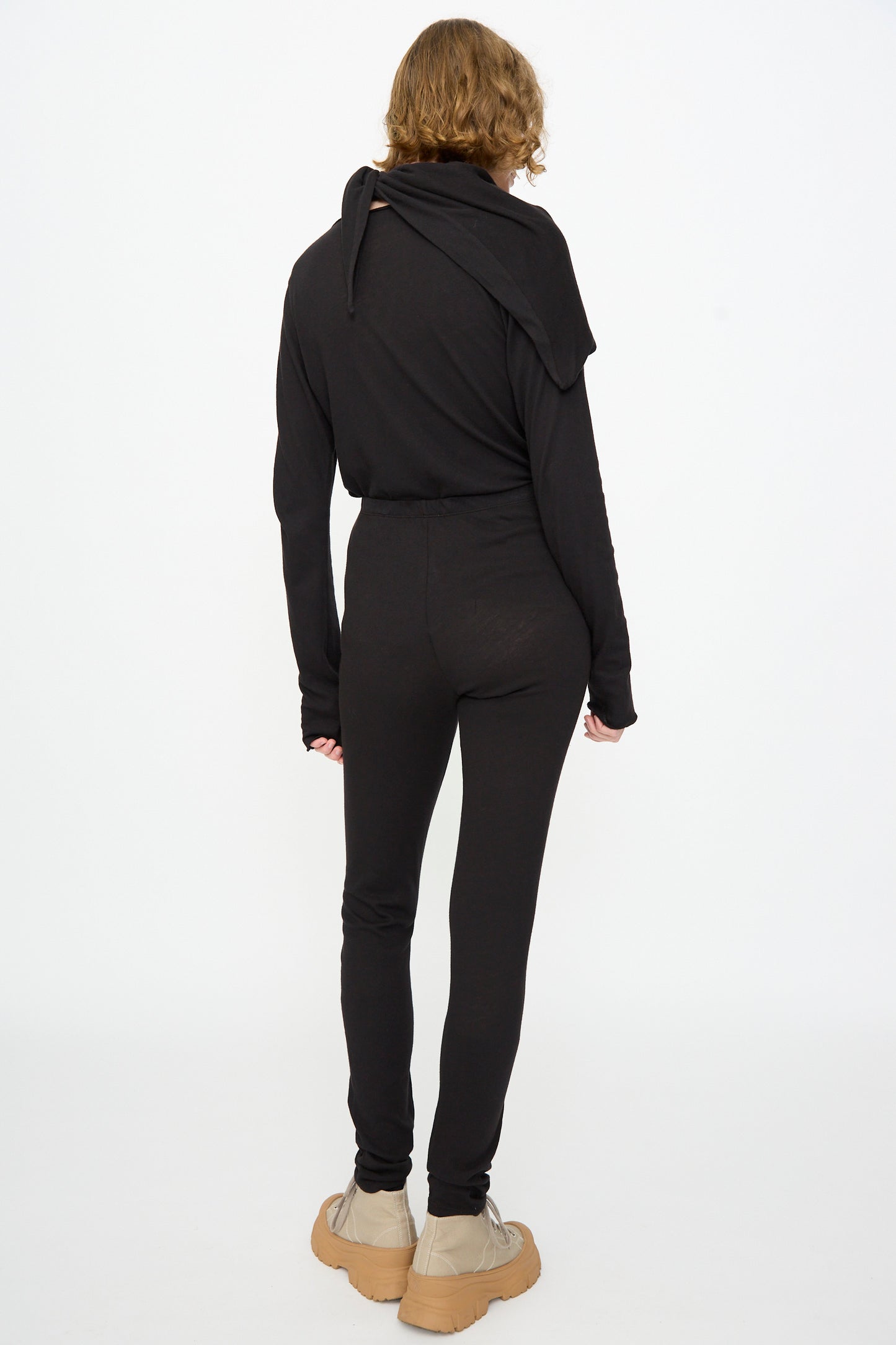 A person stands with their back to the camera, wearing Black Crane’s Leggings in Black, a black long-sleeve top, tan shoes, and a scarf draped over their shoulders.