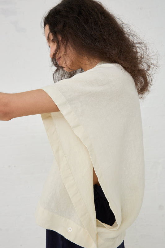 A woman is seen from behind, putting on a Black Crane Linen Origami Top in Cream with a boxy fit over a dark top.