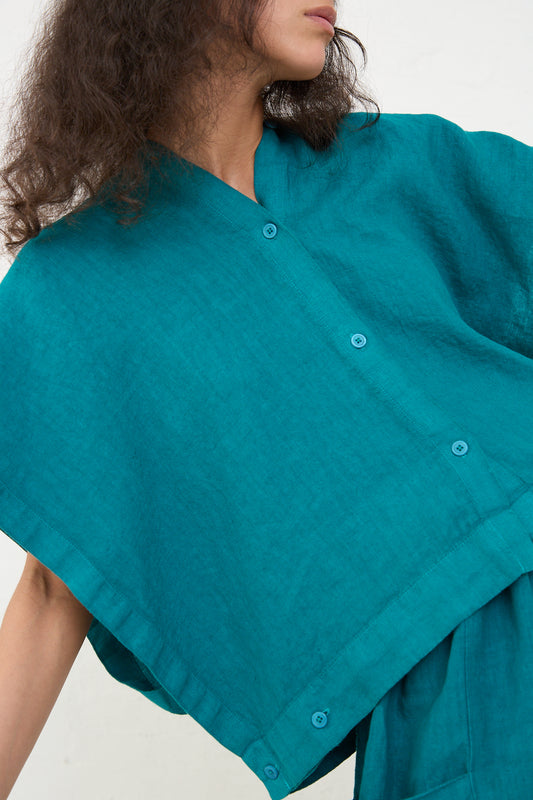 A person wearing a loose-fitting Linen Origami Top in Peacock by Black Crane, with button details on the shoulder, shown in a close-up view emphasizing the garment's texture and color. The individual's face is partially visible.