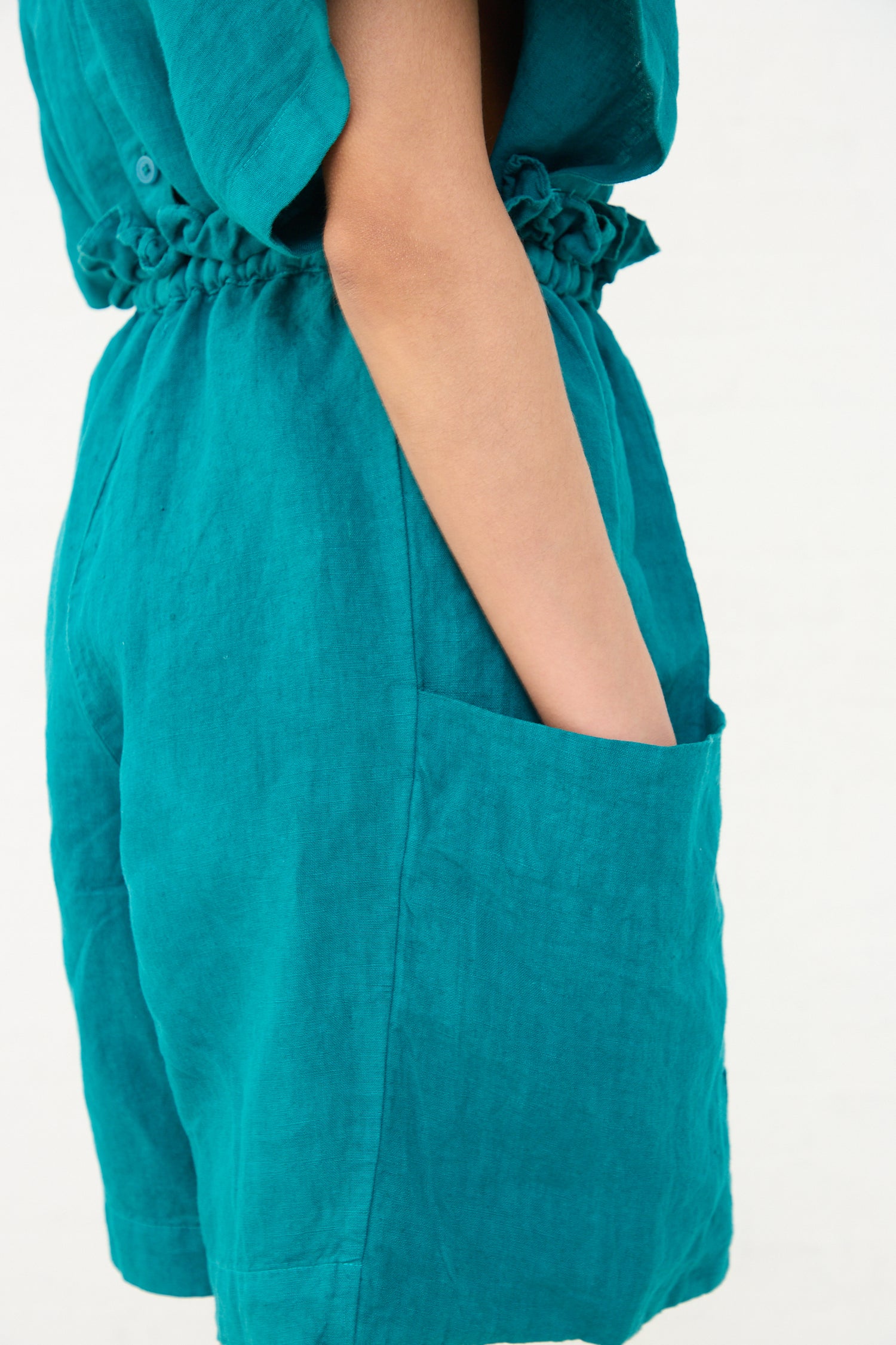 A person wearing a teal Black Crane linen dress with a hand casually tucked into the side pocket of their Black Crane parachute shorts.