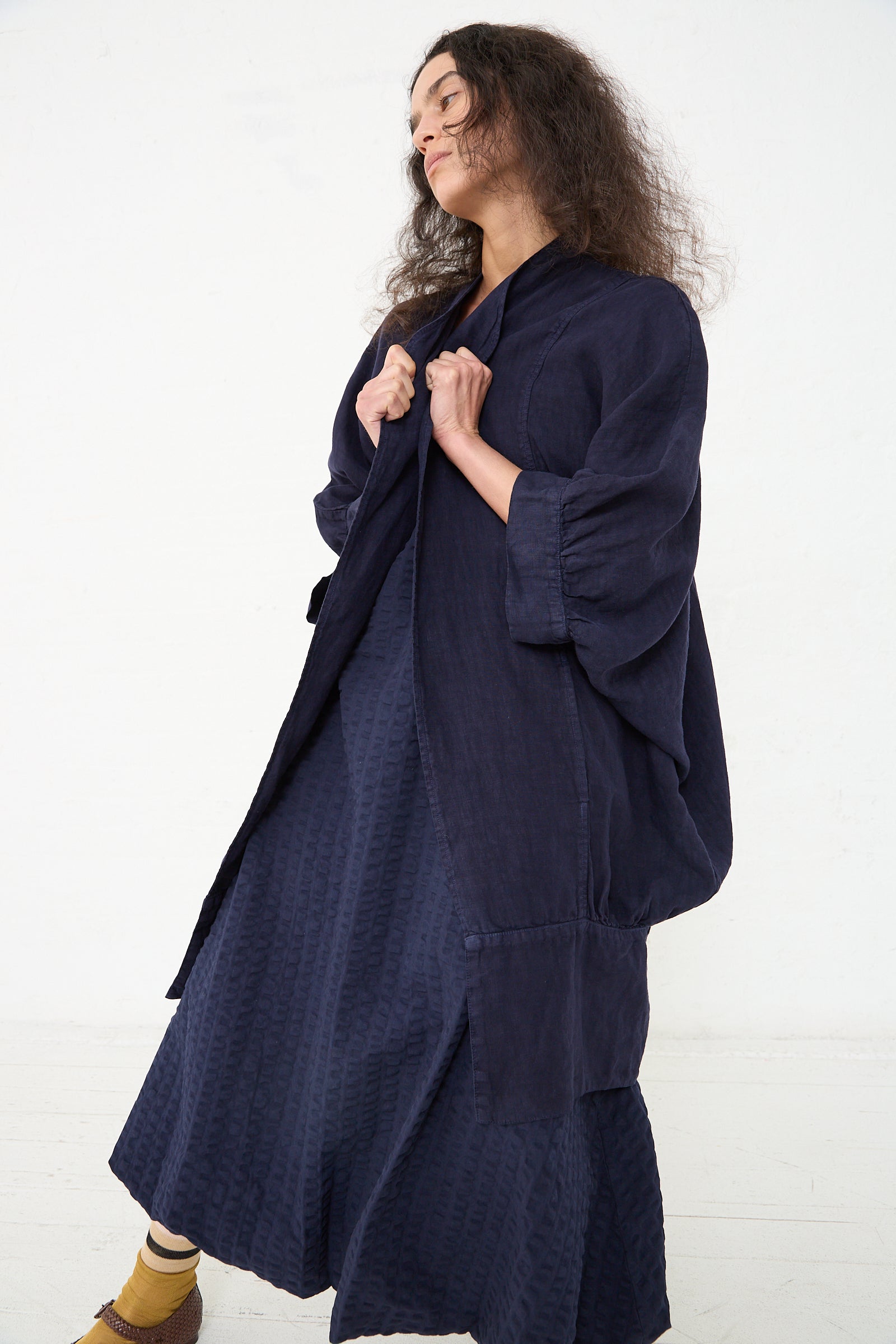 A woman in a textured navy blue ensemble consisting of a Black Crane Linen Spoon Jacket in Indigo and midi dress, paired with brown ankle socks and sandals, stands against a white background with a relaxed pose, her gaze directed slightly.