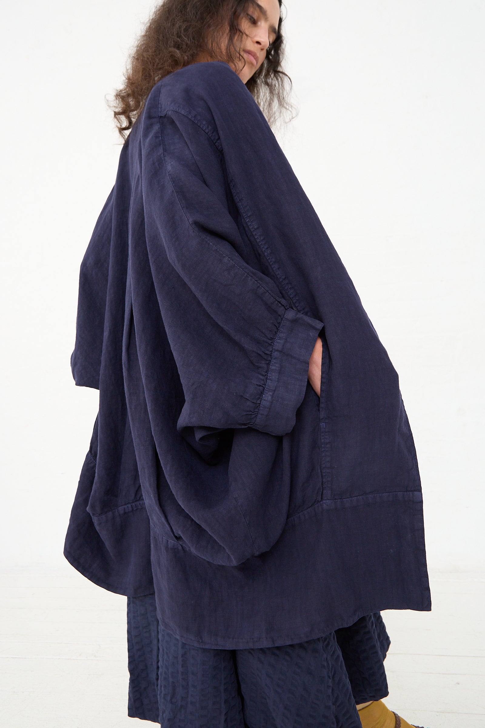 A person standing sideways in Los Angeles, wearing an oversized Black Crane Linen Spoon Jacket in Indigo with a crinkled texture, with their right hand tucked into a pocket.