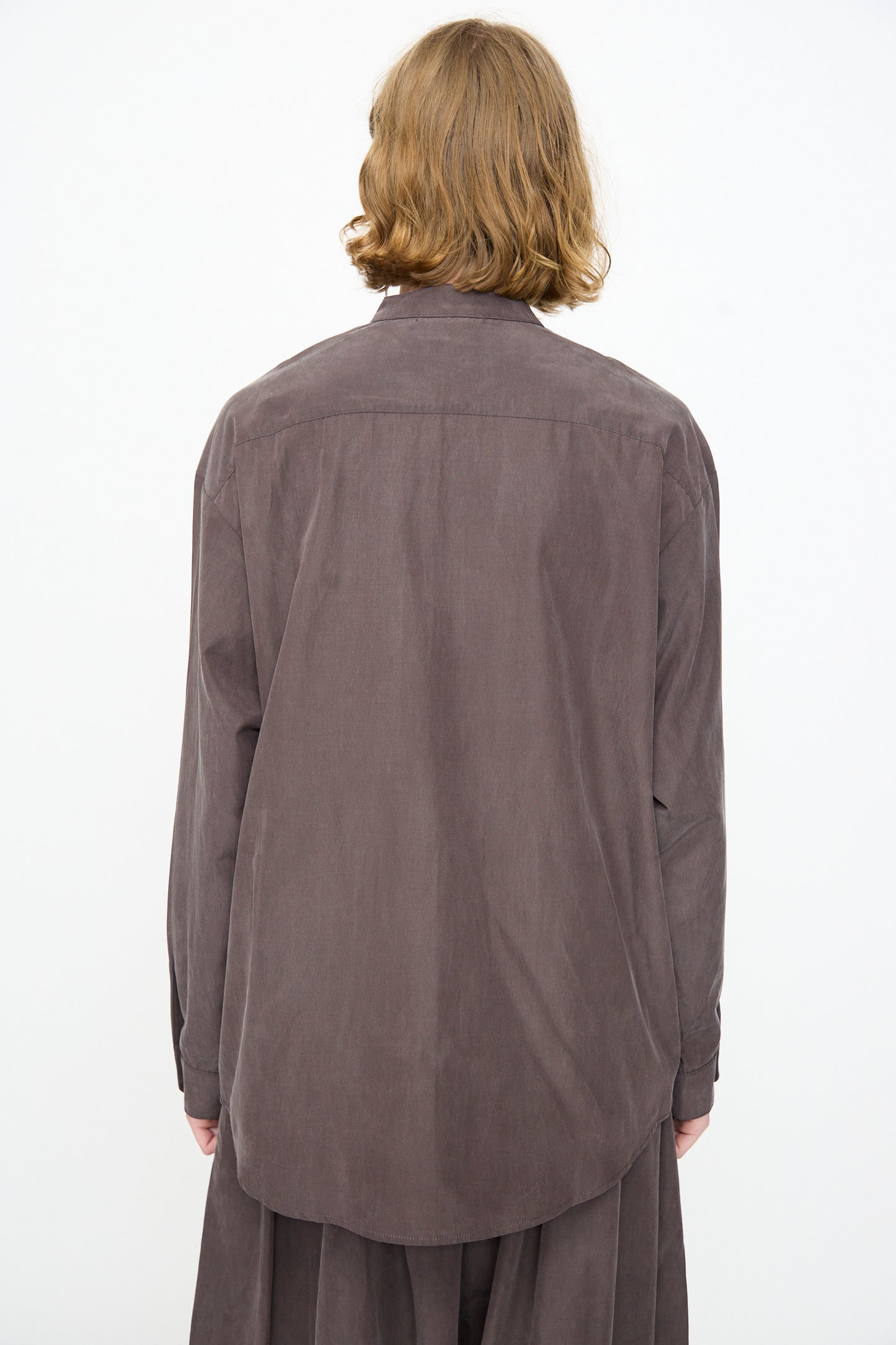 A person with shoulder-length light brown hair is seen from the back against a plain white background, wearing the Black Crane Mao Collar Shirt in Dark Plum—a loose-fitting, long-sleeve button-up crafted from a soft cotton cupro blend and featuring a band collar.