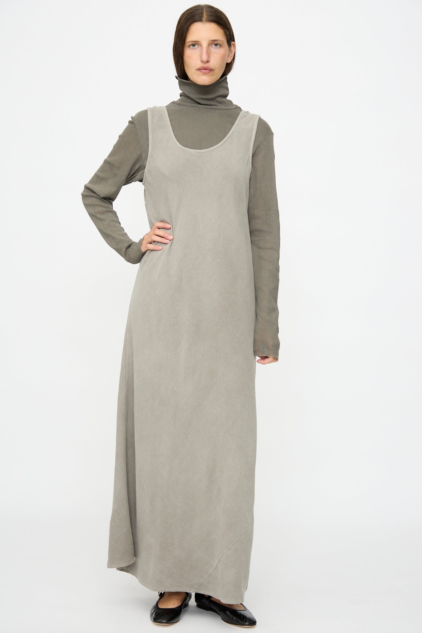 A person wears the Black Crane Maxi Dress in Dark Stone, a relaxed-fit, sleeveless style, layered over a high-neck long-sleeve top with black shoes, standing against a plain white background.