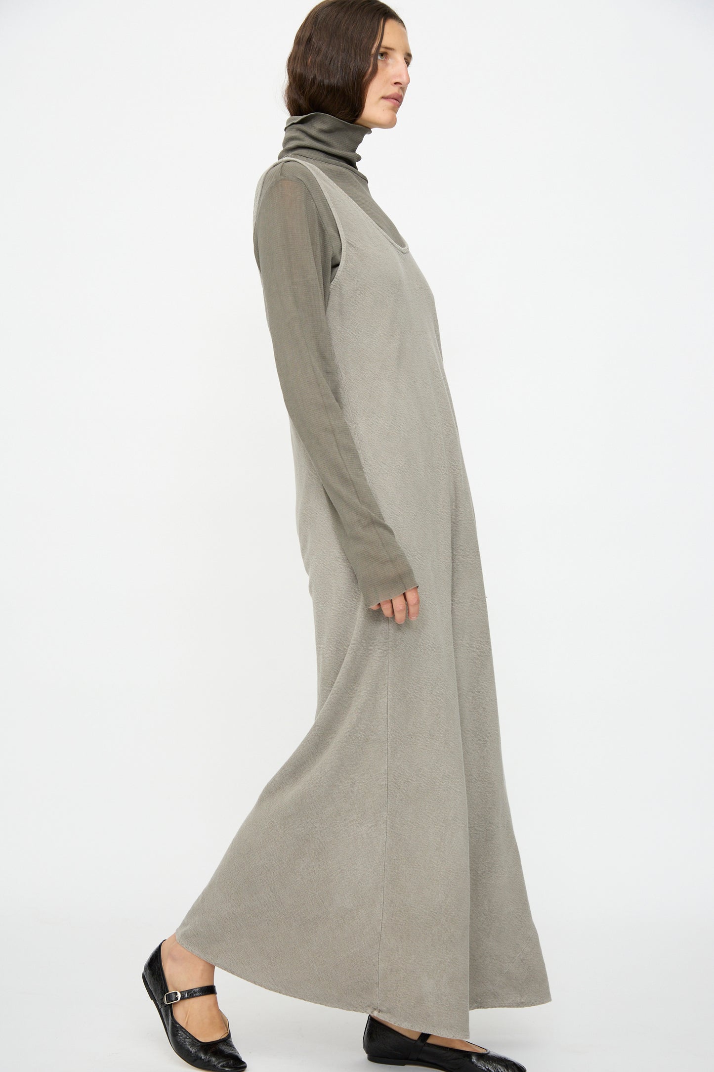 A person in the Black Crane Maxi Dress in Dark Stone, layered over a taupe turtleneck and paired with black Mary Jane shoes, stands against a plain white background.