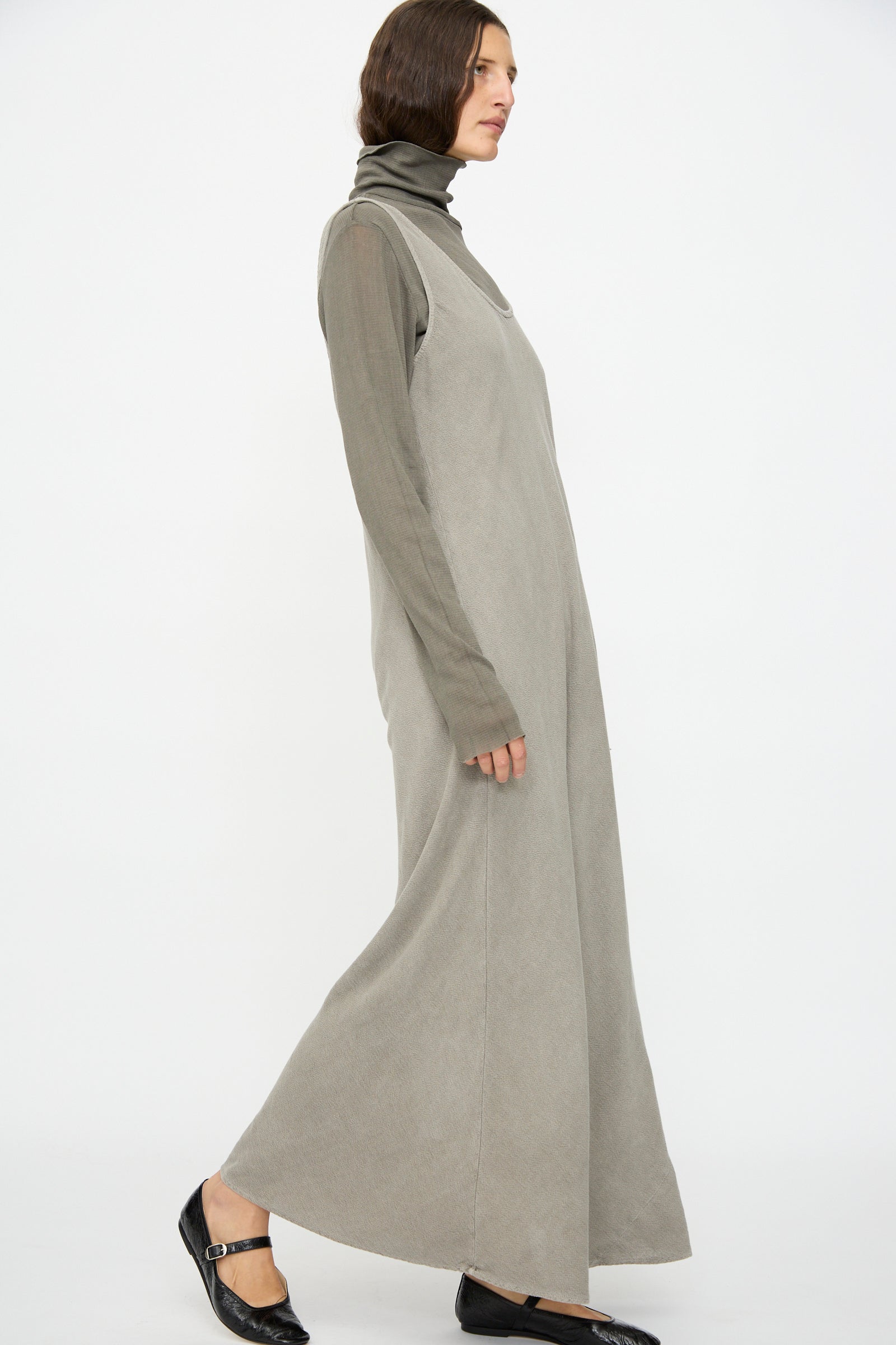 A person in the Black Crane Maxi Dress in Dark Stone, layered over a taupe turtleneck and paired with black Mary Jane shoes, stands against a plain white background.