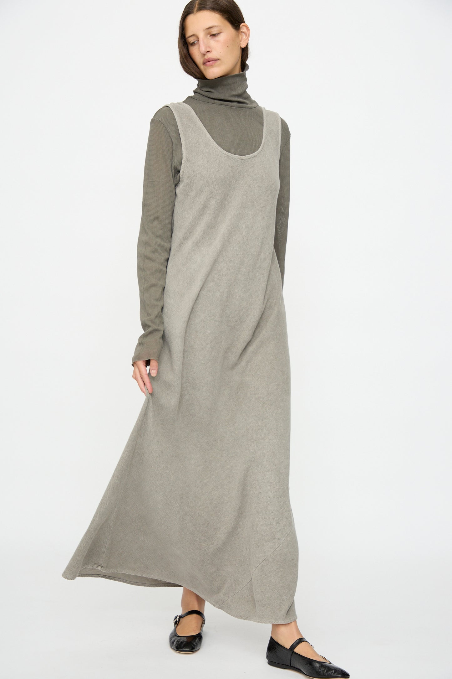 A person wears the Black Crane Maxi Dress in Dark Stone, a relaxed-fit, sleeveless style over a matching turtleneck, paired with black ballet flats, standing against a plain white background.