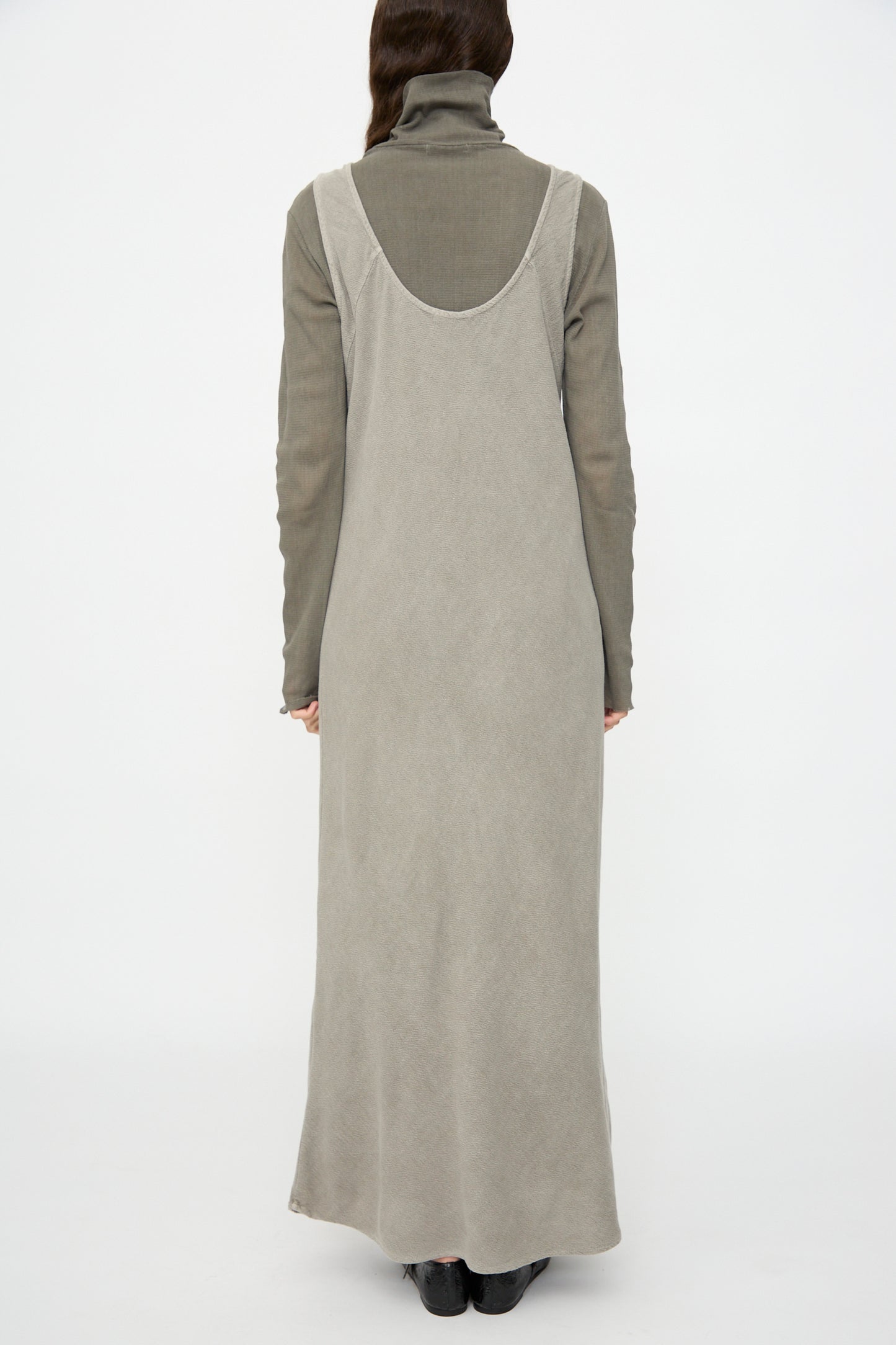 A person stands facing away, wearing the Black Crane Maxi Dress in Dark Stone—a relaxed fit, sleeveless maxi—layered over a gray long-sleeved turtleneck with black shoes, set against a plain white background.
