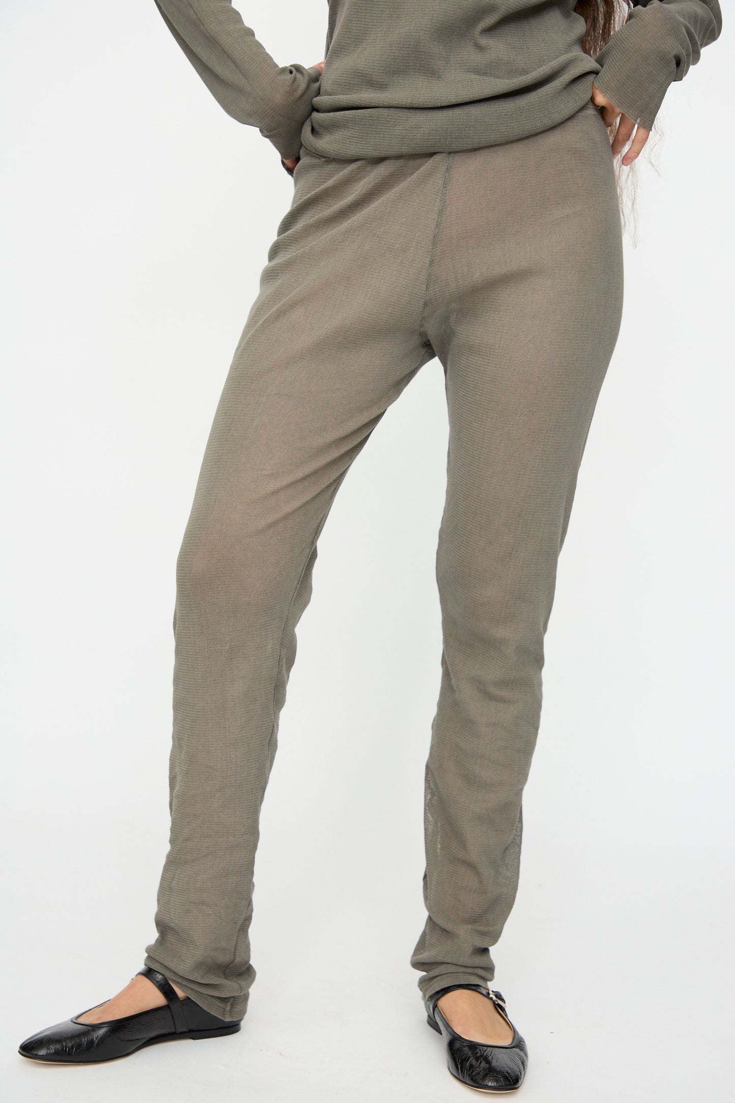 A person wears Black Crane’s Mesh Leggings in Dark Stone with an olive green slim-fit long-sleeve top and black flats, standing against a plain white background.