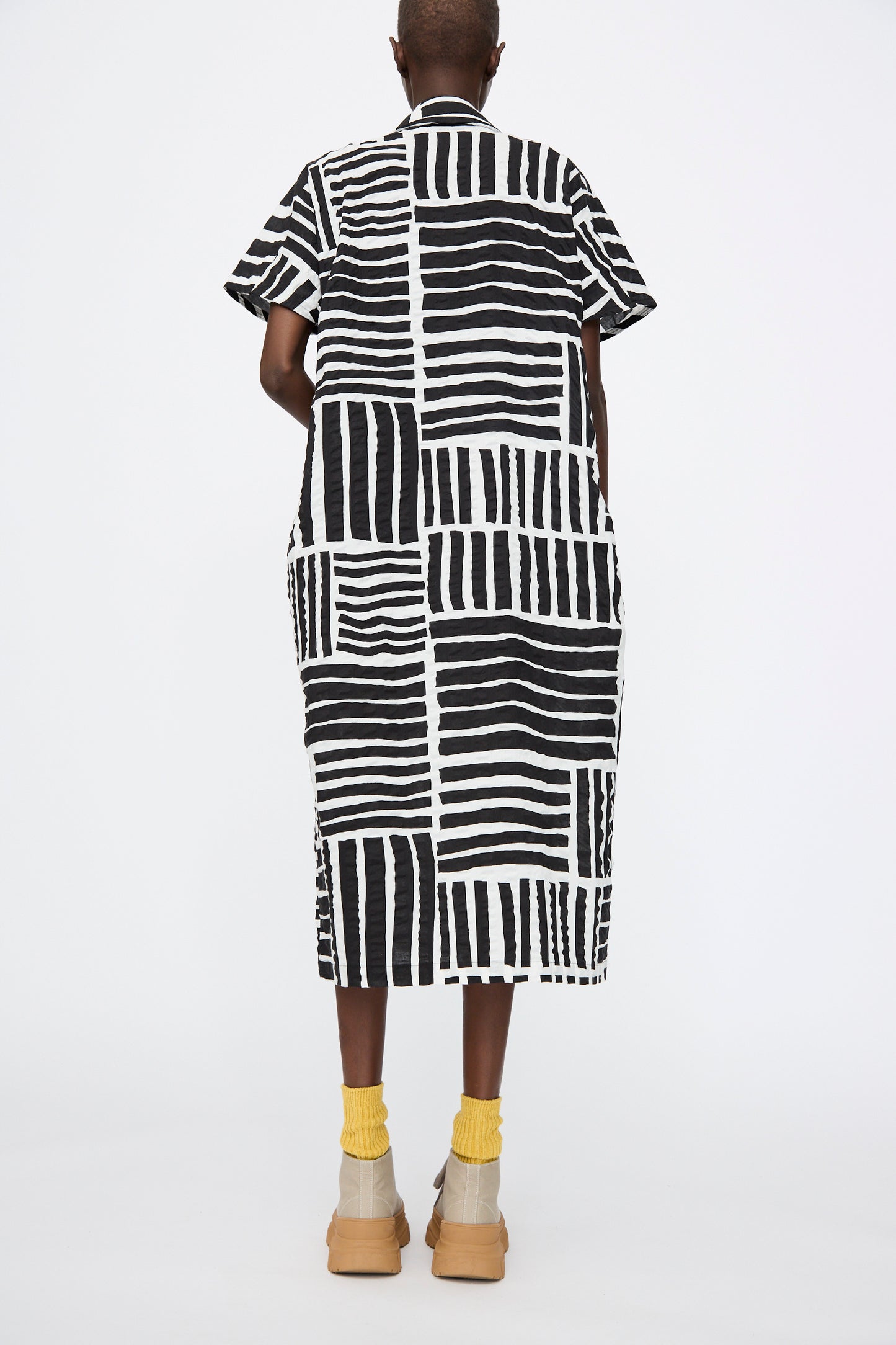 A person stands facing away against a white background, wearing the Black Crane Narrow Kite Dress in Black Print, styled with yellow socks and beige shoes.