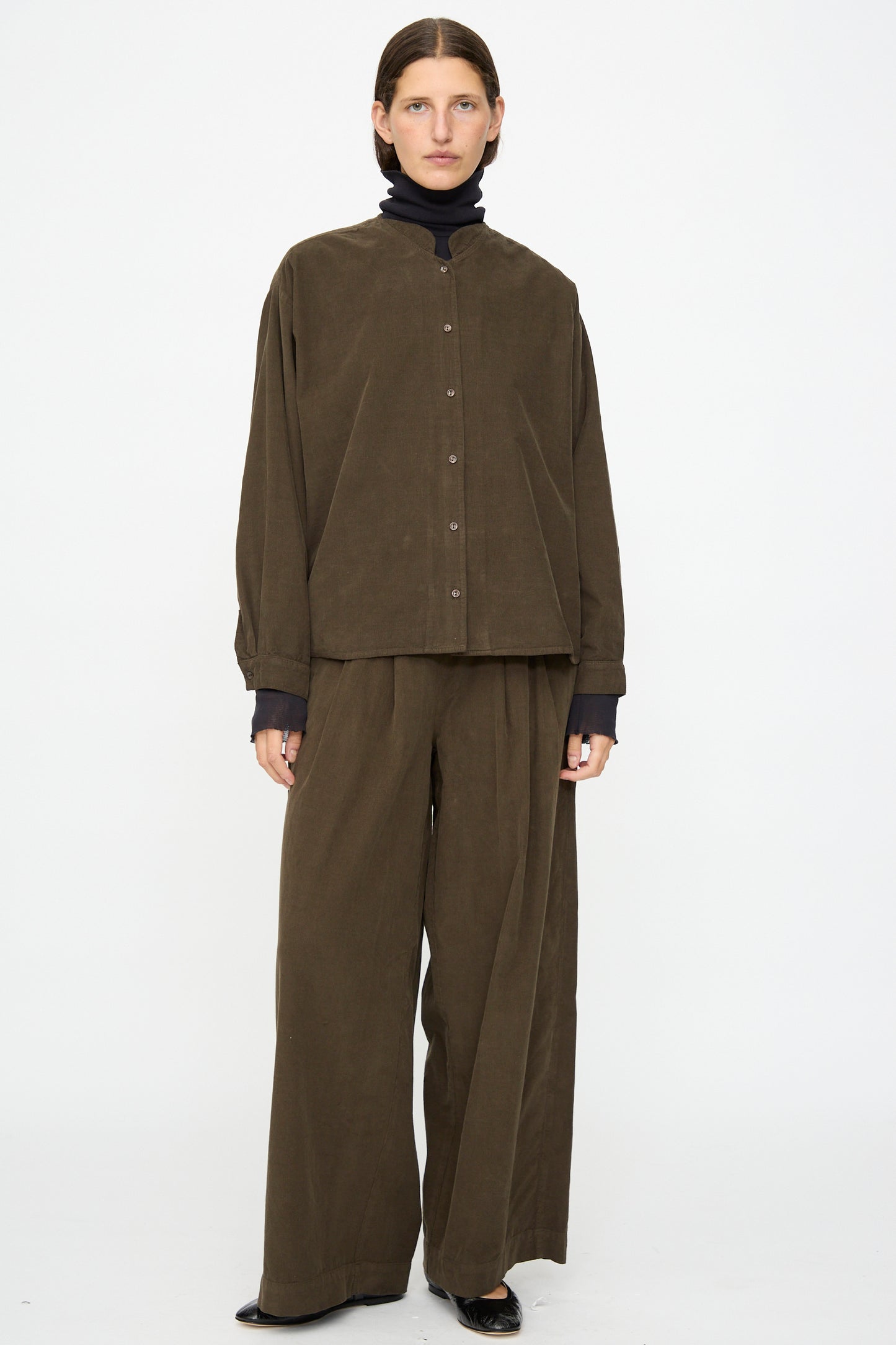 A person faces forward against a plain white background, wearing Black Crane’s Nehru Shirt in Dark Forest with matching wide-leg pants over a black turtleneck and black shoes.