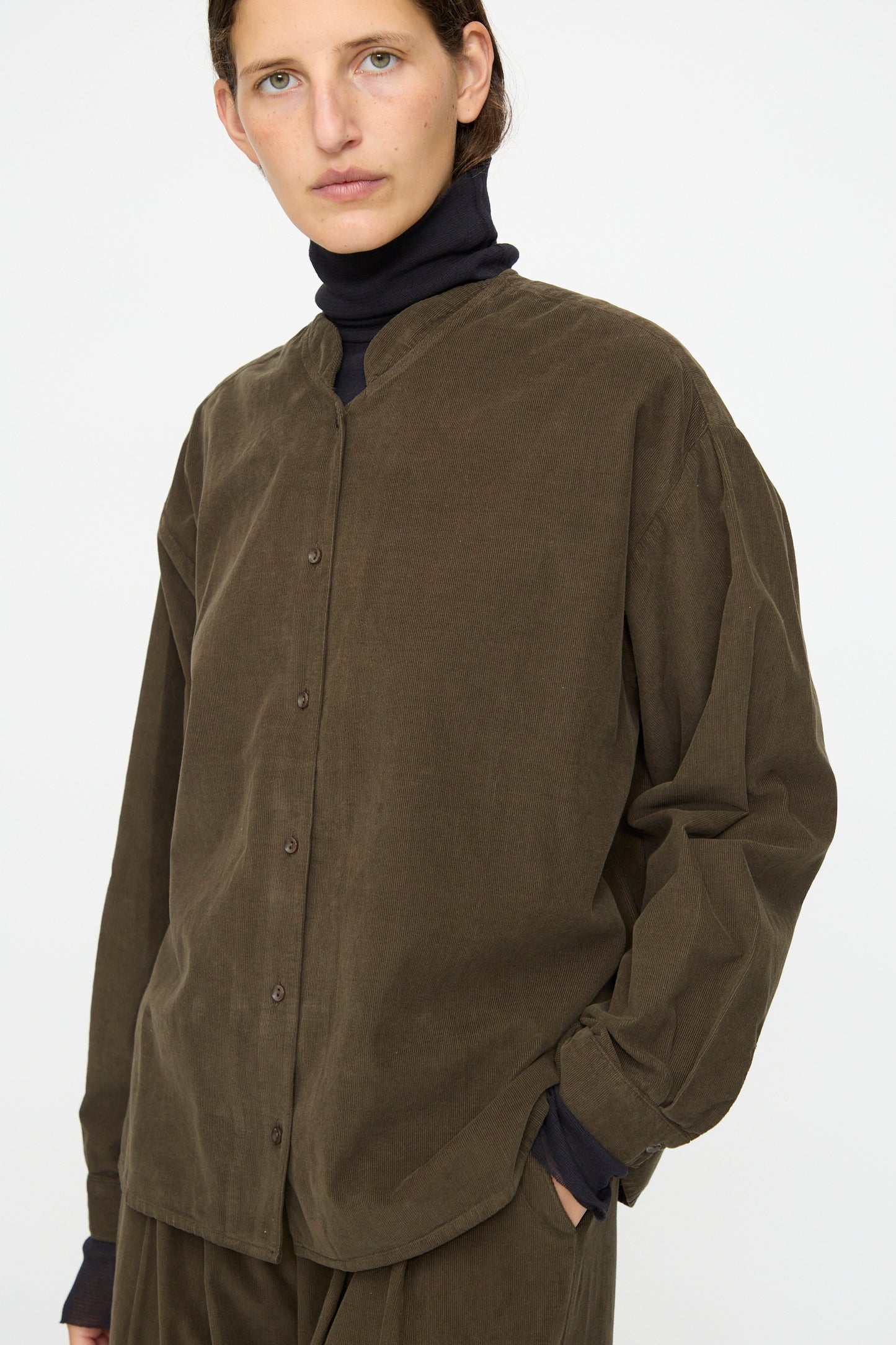 A person stands against a plain background, wearing the Black Crane Nehru Shirt in Dark Forest over a black turtleneck, with one hand in their pocket.