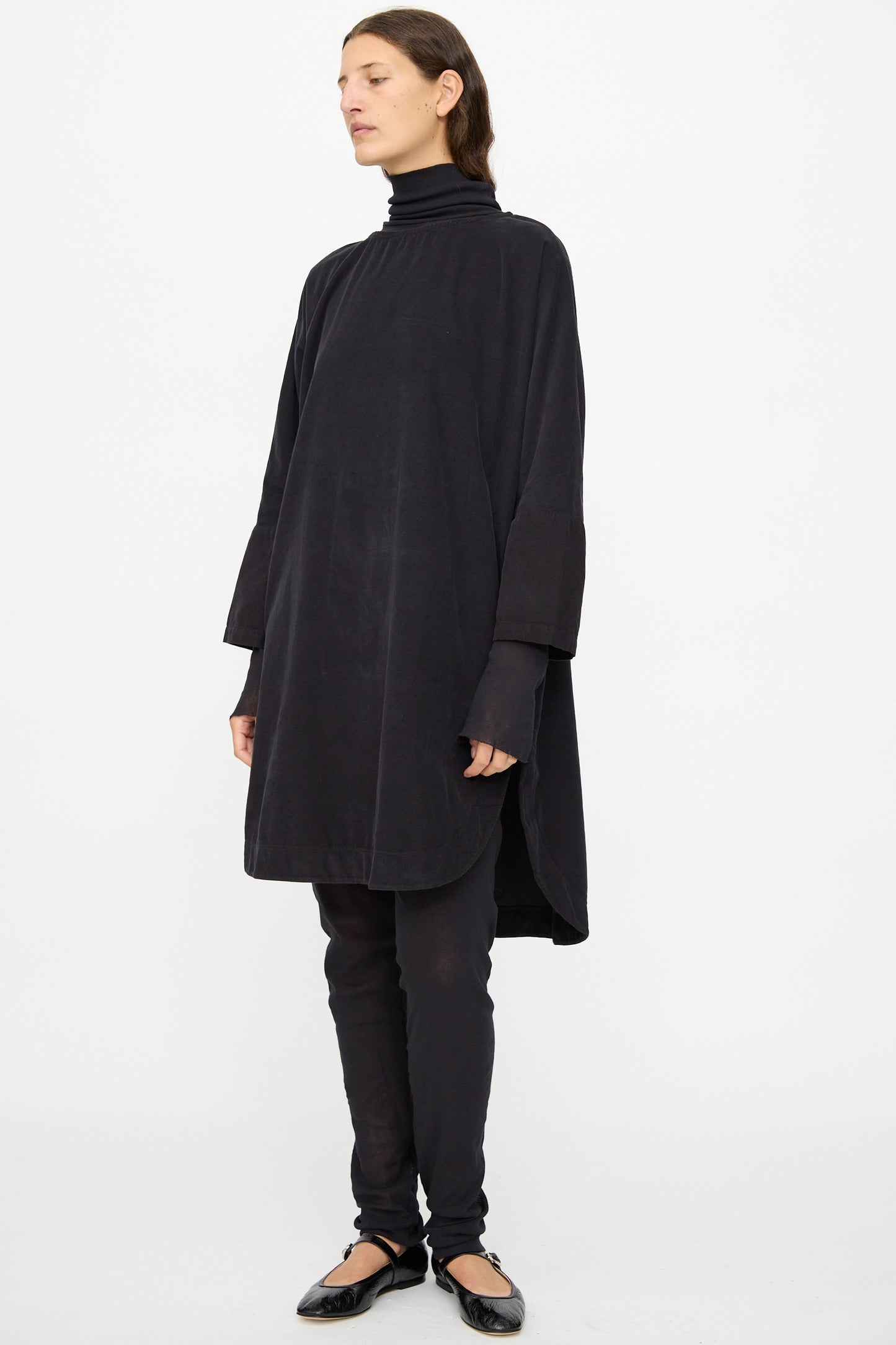 A person stands against a plain white background wearing the Black Crane Painter Dress in Dark Navy over black leggings and a black turtleneck, paired with black ballet flats.