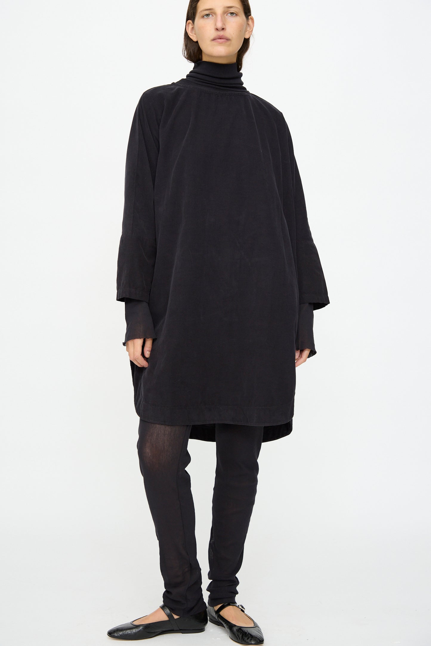 A person stands against a plain background wearing the Black Crane Painter Dress in Dark Navy, made in the USA, layered over a black turtleneck, black leggings, and black ballet flats.