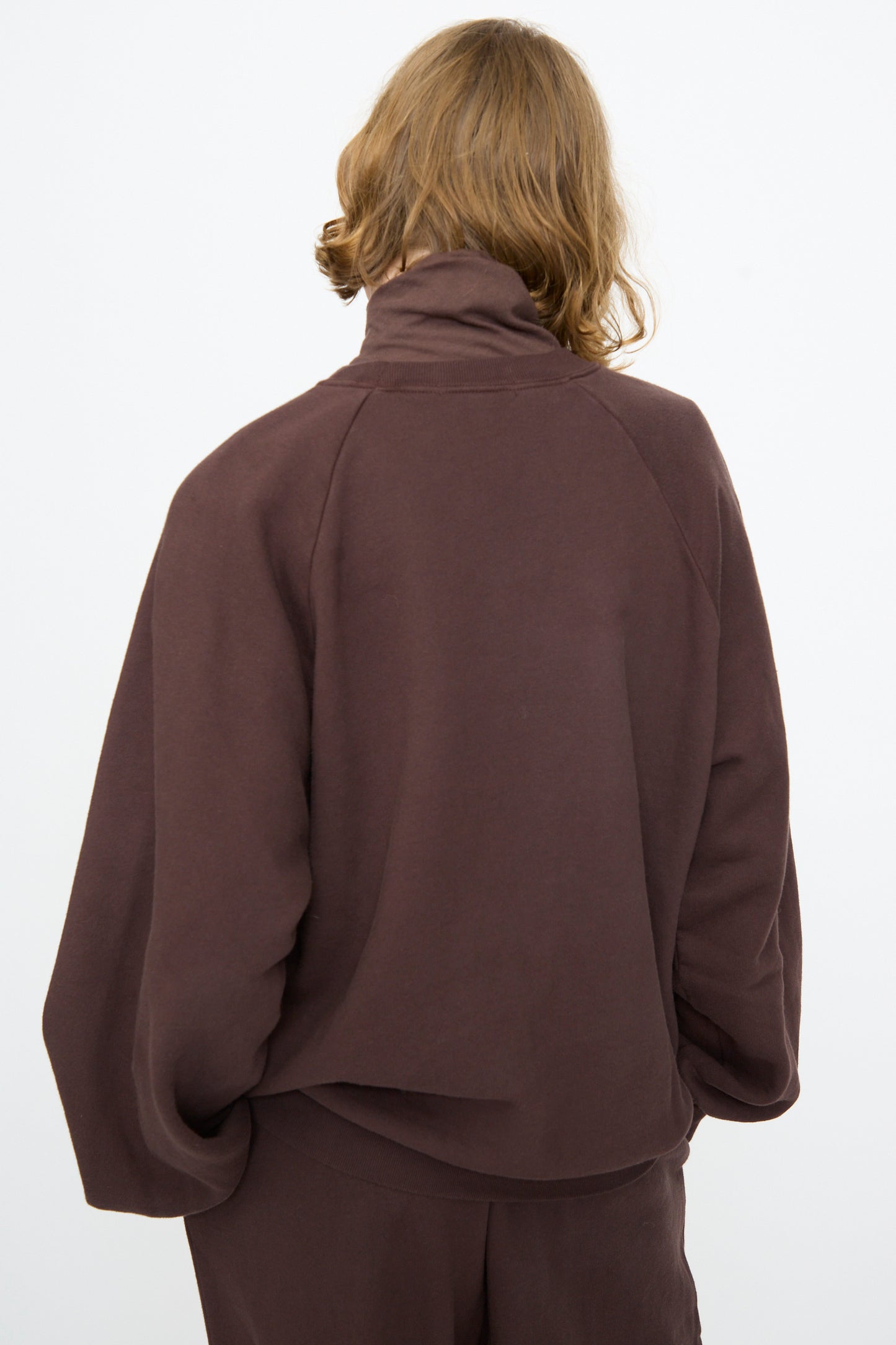 A person with shoulder-length light brown hair is seen from behind, wearing the Black Crane Raglan Sweatshirt in Dark Cacao paired with matching pants, standing against a plain white background.