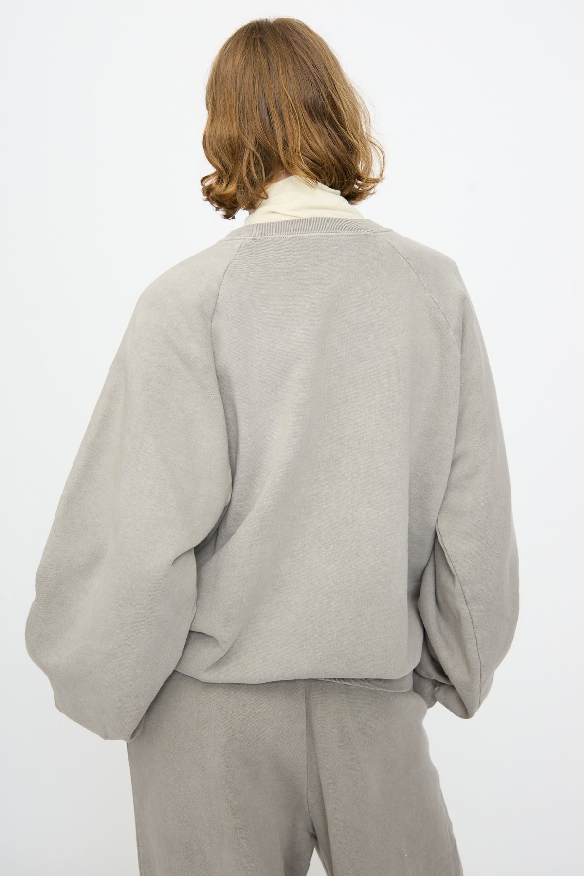 A person with shoulder-length light brown hair is shown from the back against a plain white background, wearing the Black Crane Raglan Sweatshirt in Stone—a loose, organic cotton grey pullover—paired with matching pants.