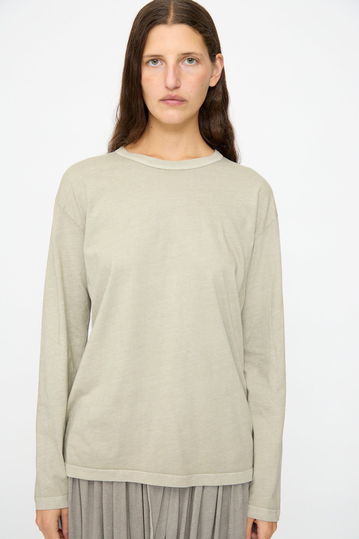 A person with long brown hair wears the Black Crane Regular Long Sleeve Tee in Limestone and a gray pleated skirt, standing against a plain white background.