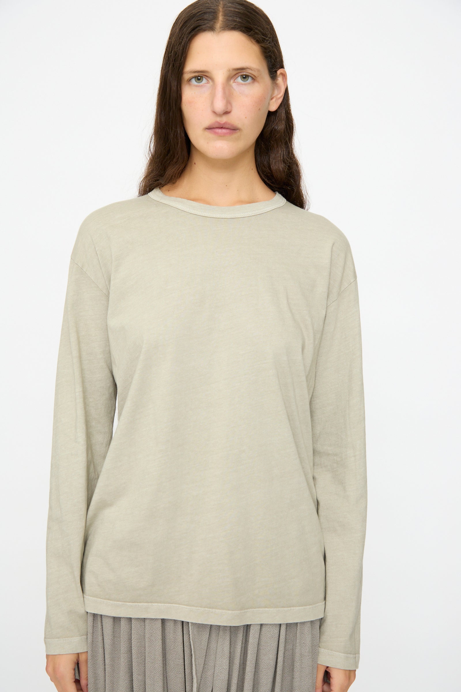 A person with long brown hair wears the Black Crane Regular Long Sleeve Tee in Limestone and a gray pleated skirt, standing against a plain white background.