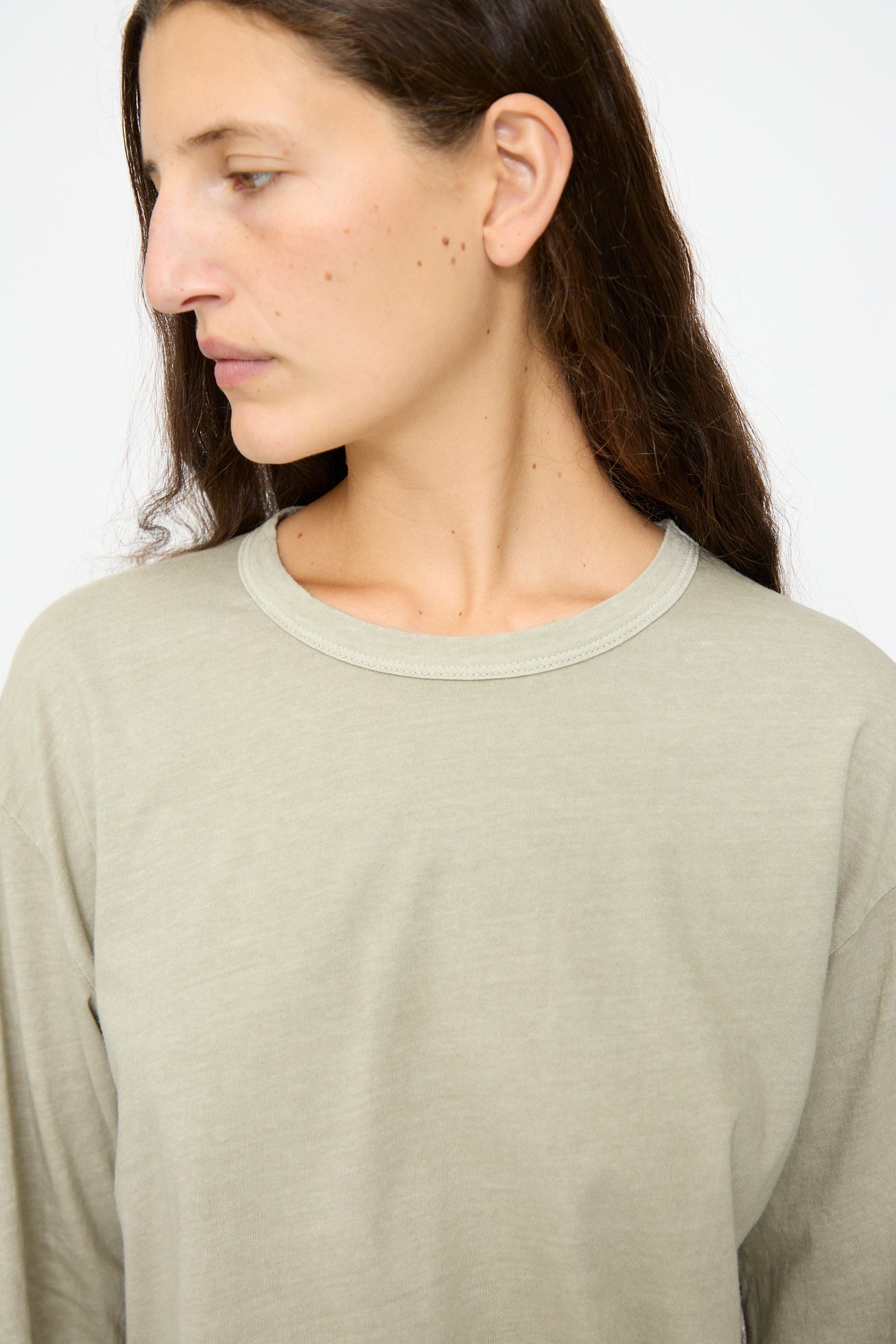 A person with long brown hair wears the Black Crane Regular Long Sleeve Tee in Limestone and looks to the left against a plain background.