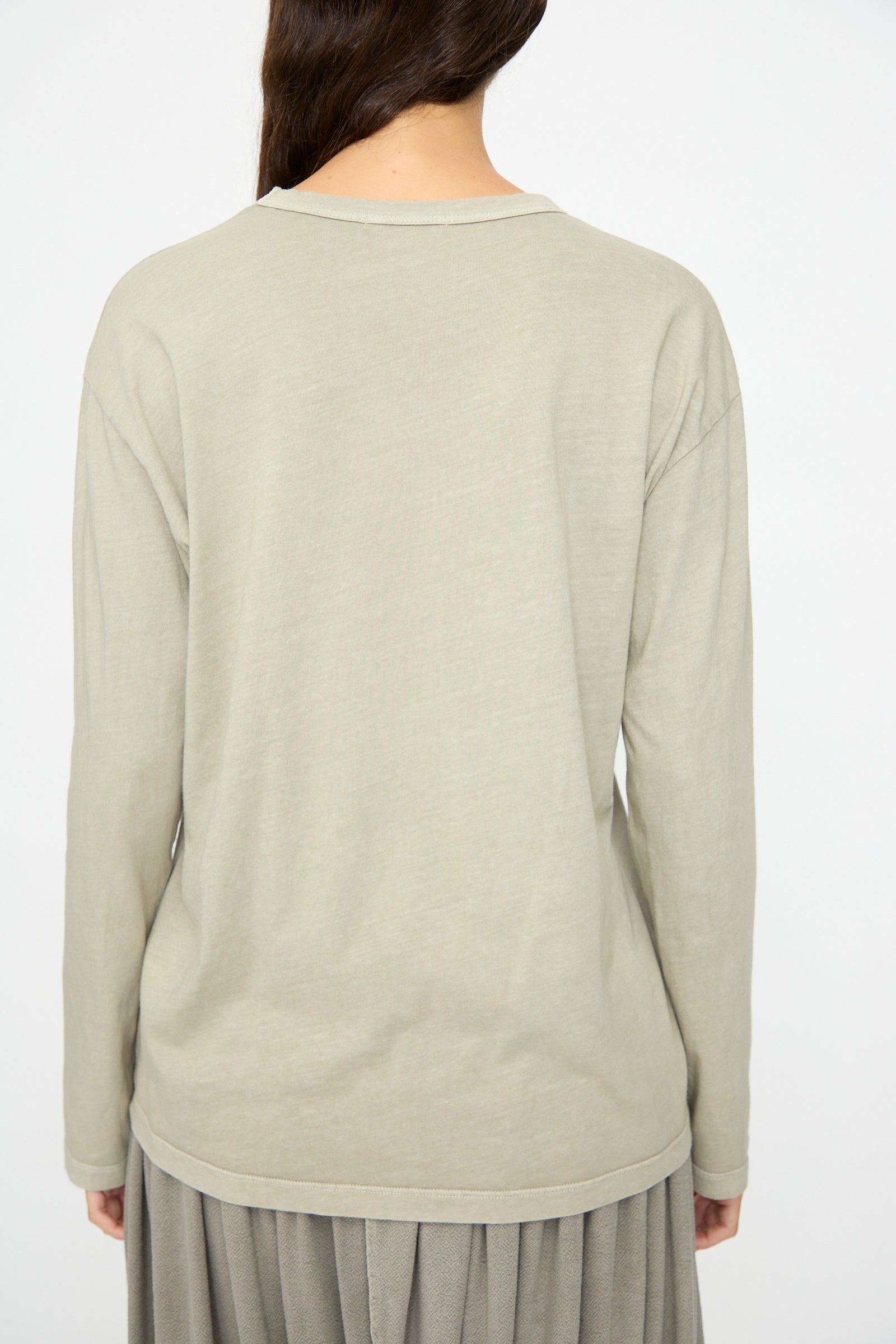 A person with long brown hair, seen from behind, wears the Black Crane Regular Long Sleeve Tee in Limestone and a gray pleated skirt against a plain background.