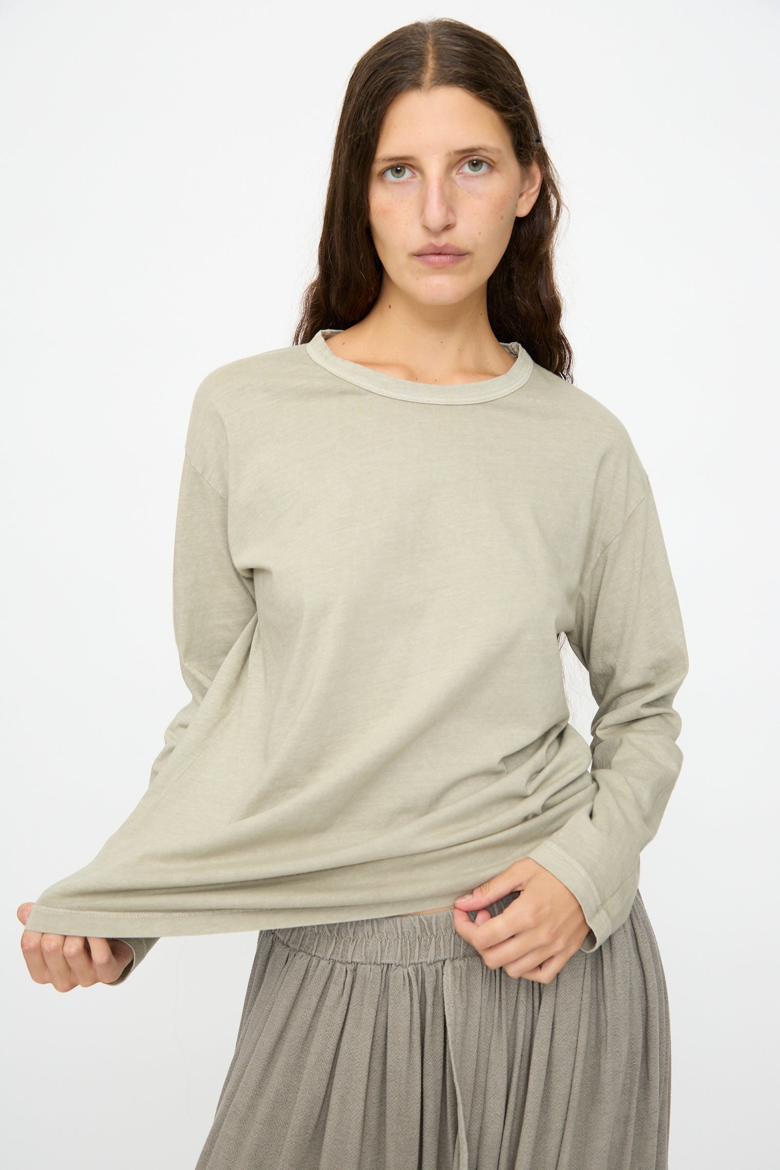 A person with long brown hair wears the Black Crane Regular Long Sleeve Tee in Limestone, paired with a gray skirt, standing against a plain white background while holding the side of their shirt.
