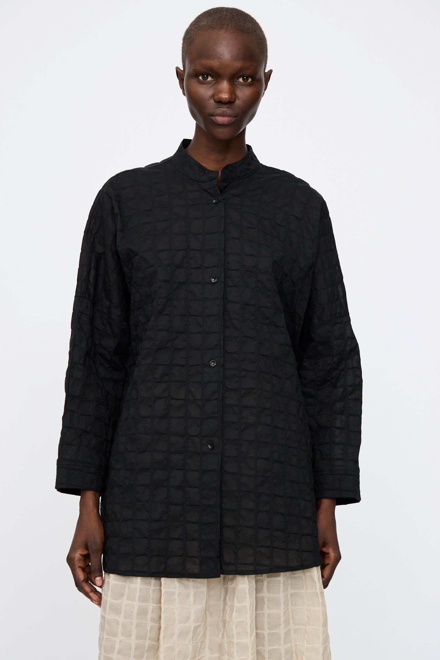 A person stands facing forward, wearing the Black Crane Ripple Finish Shirt in Black—a textured Japanese cotton button-up with long sleeves and a band collar—paired with a light-colored skirt.