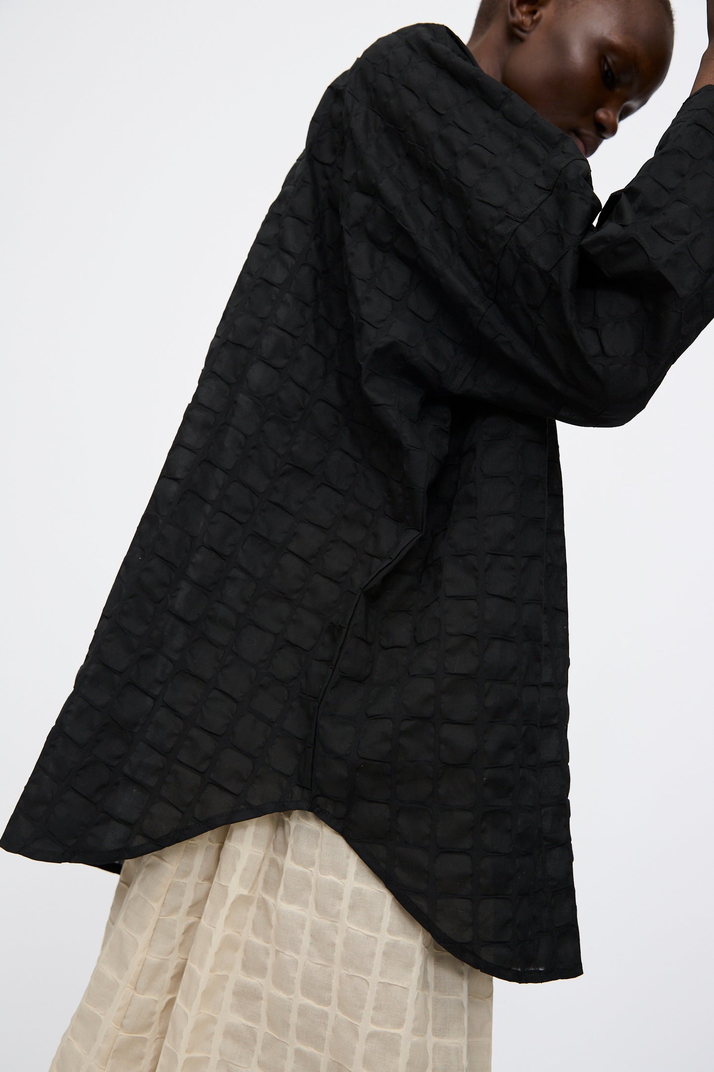 Person wearing the Ripple Finish Shirt in Black by Black Crane, styled oversized with a band collar over a light, textured skirt, standing against a plain light background.