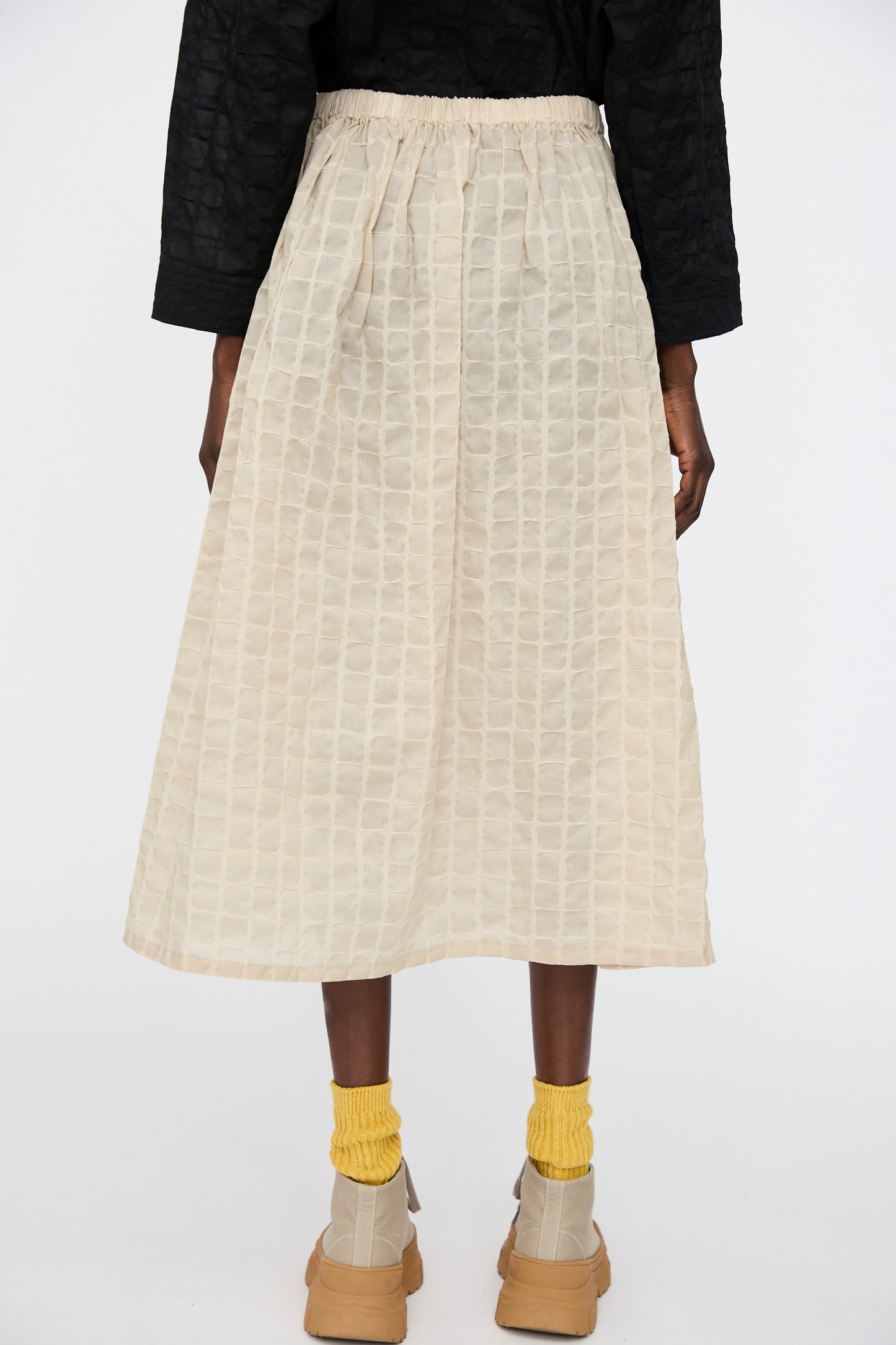A person faces away, wearing the Black Crane Ripple Finish Skirt in Beige—a Japanese cotton textured midi with a subtle check pattern—paired with a black top, beige shoes, and yellow socks against a plain white background.