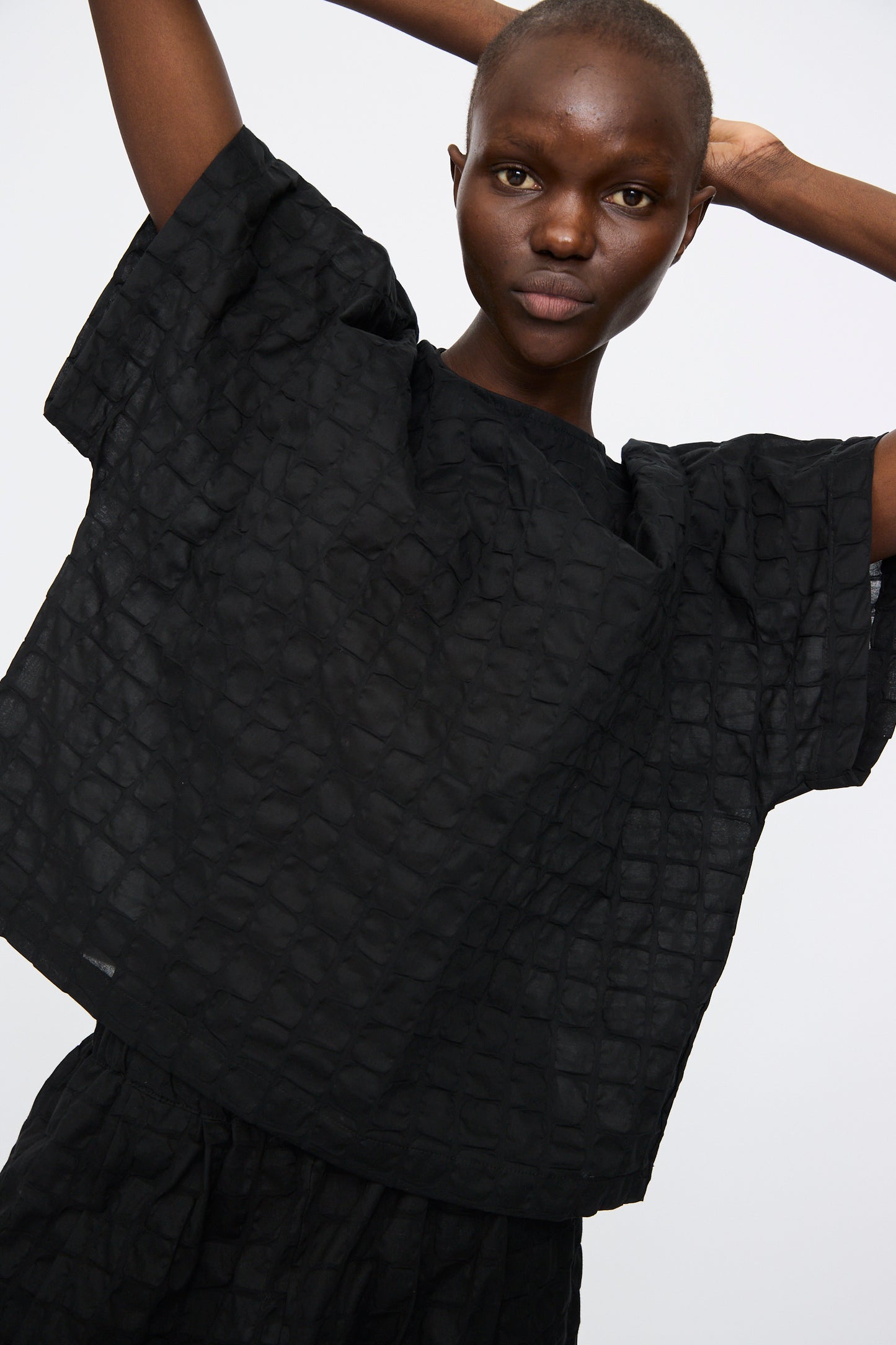 Person wearing the Black Crane Ripple Finish Top in Black—a loose, boxy Japanese cotton shirt with short sleeves and subtle texture—stands against a plain light background with arms raised and crossed behind their head.