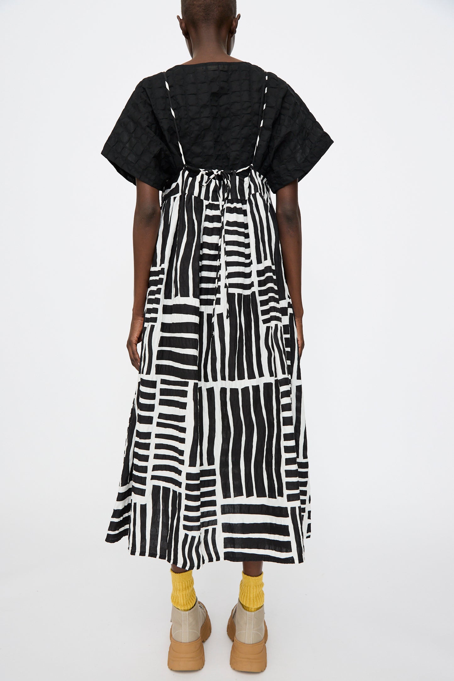 A person stands against a white background wearing the Black Crane Strap Dress in Black Print over a black textured top, paired with yellow socks and beige platform shoes. The photo captures the back view.
