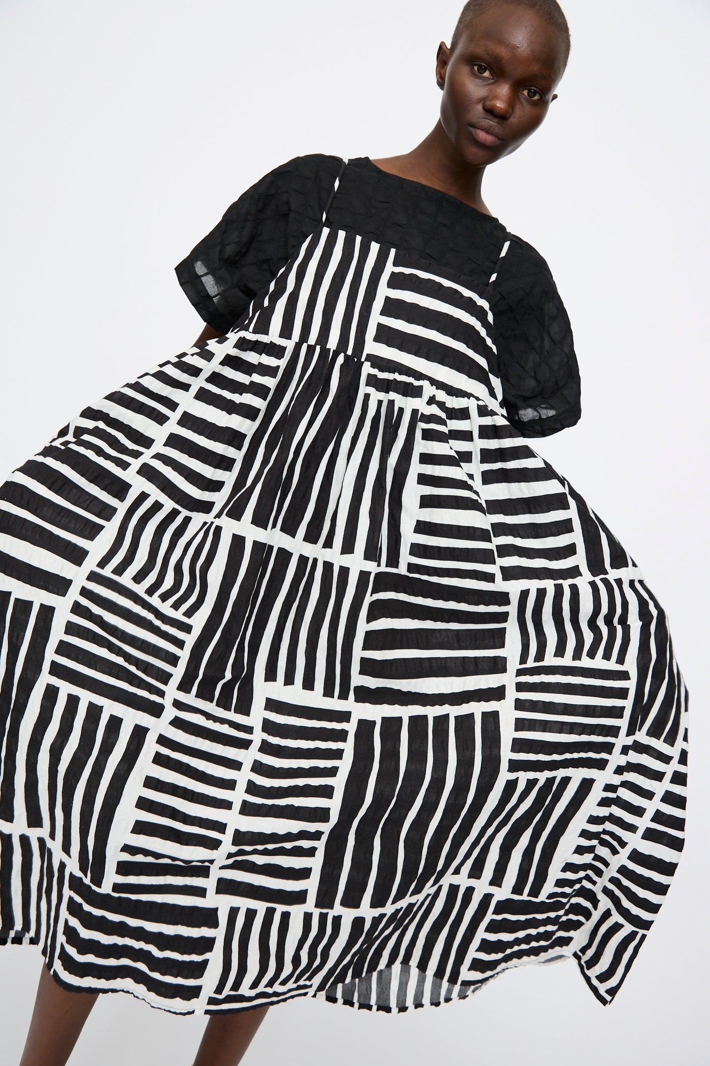 A person wears the Black Crane Strap Dress in Black Print, a loose black midi dress with bold graphic patterns, standing against a plain white background.