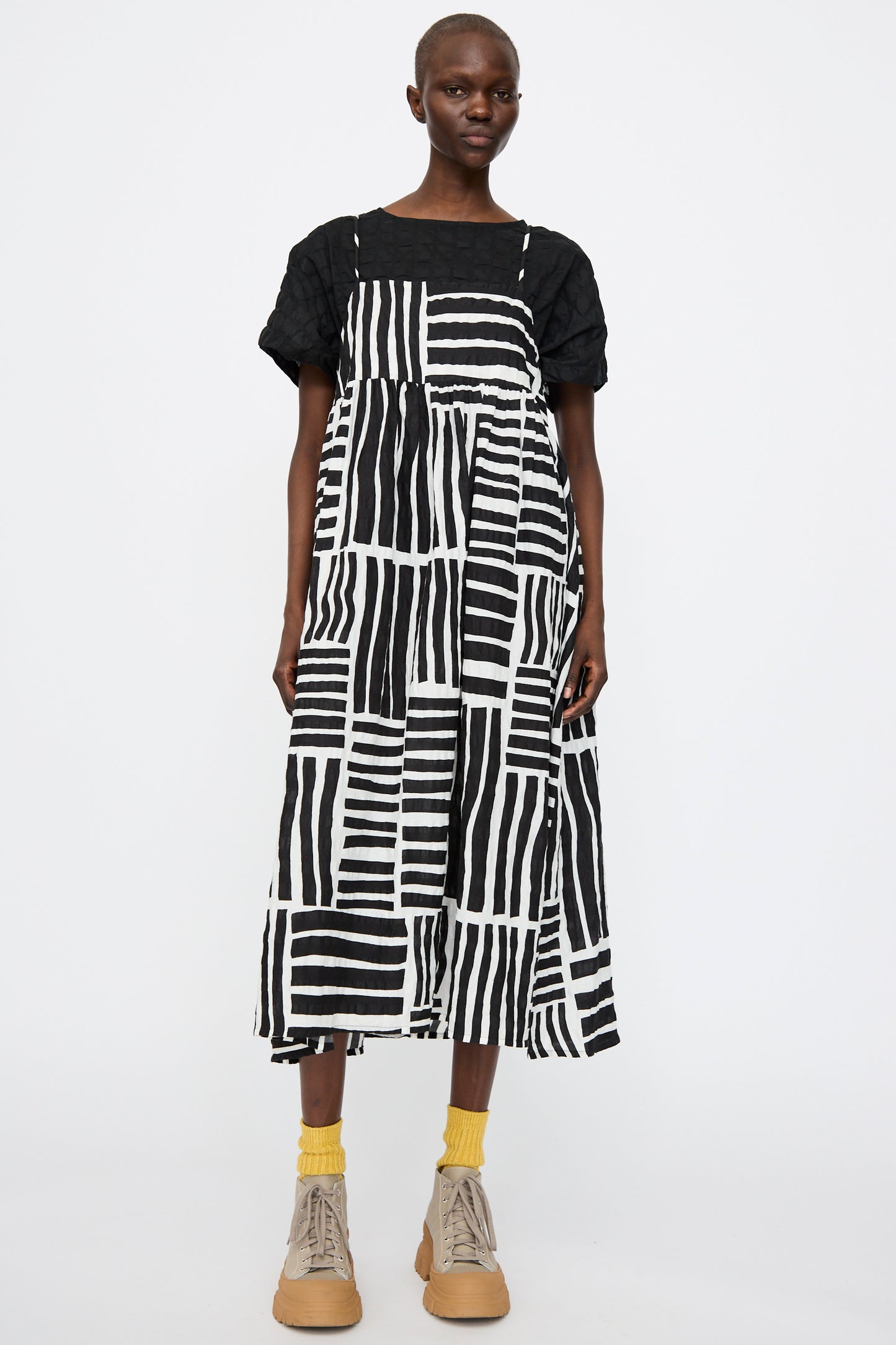 Model wearing the Black Crane Strap Dress in Black Print over a black top, paired with yellow socks and beige boots, stands against a plain white background.