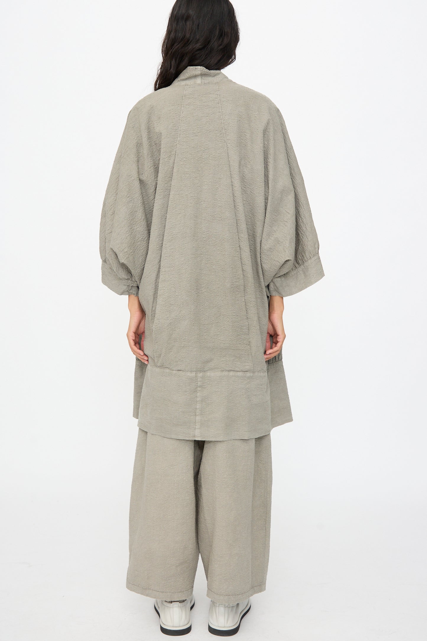 A person stands with their back to the camera, wearing the Black Crane Textured Cotton Spoon Jacket in Pale Grey—an oversized top paired with matching wide-leg pants and white shoes—set against a plain white background.