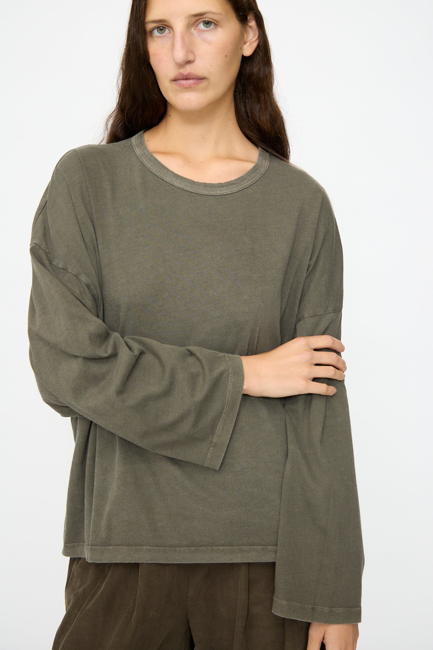 A person with long brown hair wears the Black Crane Wide Long Sleeve Tee in Slate, paired with brown pants, standing against a plain light background.